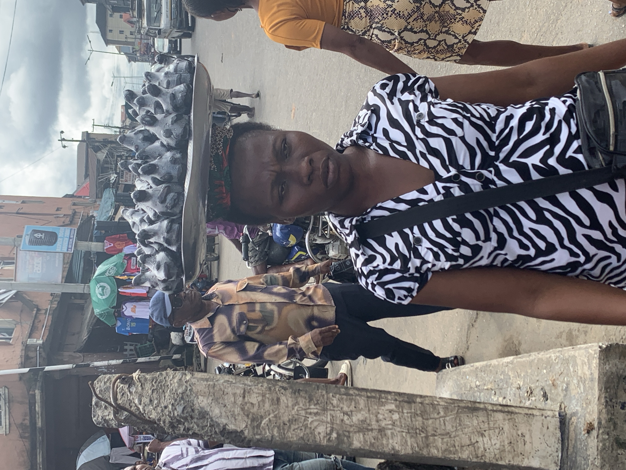 In Aba, Nigeria, a woman wearing a black and white zebra-print shirt and a patterned headwrap carries a large metal tray laden with numerous small, dark figures on her head. The figures appear to be clay figurines, possibly depicting animals, with distinct heads and bodies. The woman is positioned in the foreground, facing the camera with a neutral expression. Behind her, a man in a camouflage-patterned shirt and dark pants walks to her left, wearing a blue cap and sunglasses. To the woman's right, another person is partially visible, wearing an orange shirt and a snakeskin-patterned skirt. The background shows a bustling street scene with various shops, motorcycles, umbrellas, and other pedestrians. The overall environment suggests a vibrant market or commercial area in the city.