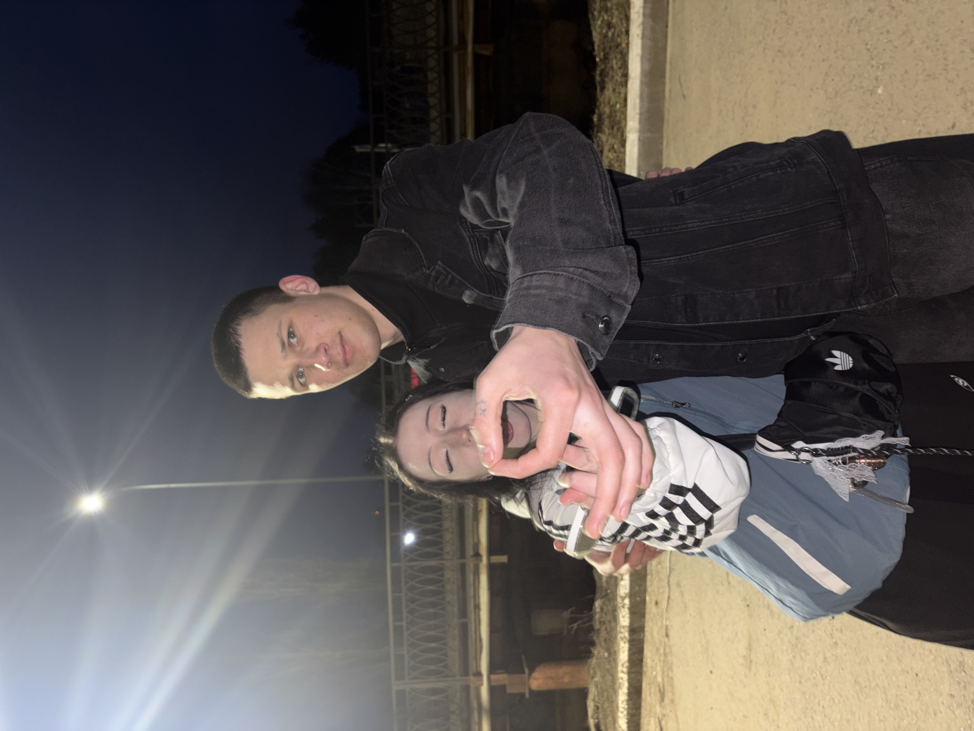 This vertically oriented image captures two young individuals, a male and a female, interacting outdoors at night in Chaikovsky, Russia. The male, wearing a dark denim jacket, stands behind the female, looking towards the camera with a neutral expression. His hand is positioned near the female's face. The female, in a light blue or grey jacket with a black and white checkered pattern on a sleeve, and dark pants, has her eyes closed and mouth slightly open in what appears to be a playful or exaggerated expression, looking up towards the male's hand. She is also wearing an item, possibly a boot or bag, adorned with the Adidas trefoil logo and white stripes. Keys dangle from her outfit.
The scene is set at night, illuminated by prominent streetlights, one of which creates a strong starburst effect on the left side of the frame. The background is dark, revealing distant trees and an industrial-style fence or railing structure, suggesting an urban or semi-urban outdoor location, possibly a path or bridge. The ground appears to be paved or concrete. The image captures a casual and friendly moment of interaction between the two subjects. Aside from the Adidas branding, no other text is visible.