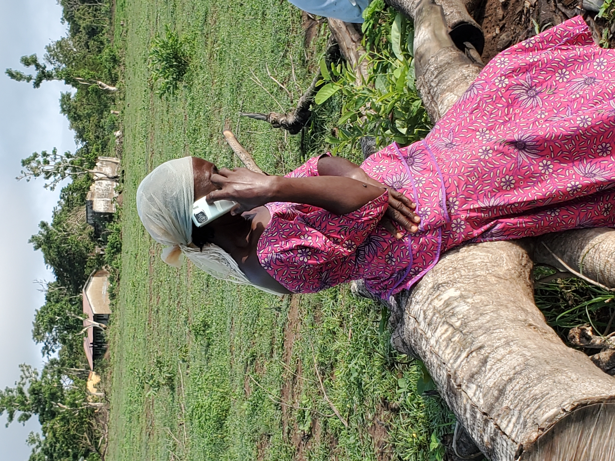 A woman sits on a fallen tree trunk in a grassy field in Mararaba, Nigeria. She is wearing a pink floral dress and a white head covering, and she is holding a white smartphone to her ear. In the background, there are trees, grass, and buildings.