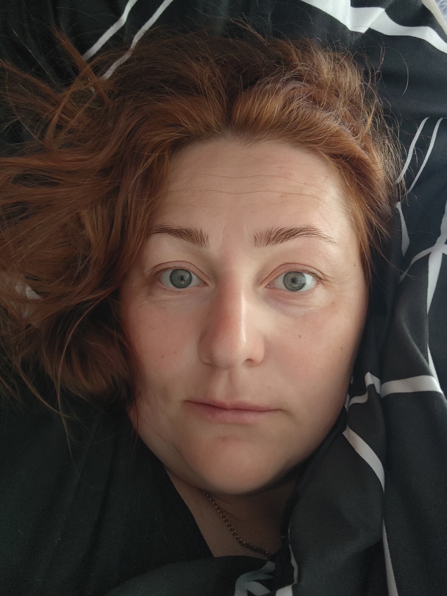 A close-up selfie shows a woman with auburn hair and light green eyes lying down against a black and white striped duvet. Her face is centered in the frame, with a neutral expression. Some wrinkles are visible on her forehead and around her eyes. A thin gold chain is visible around her neck. The background is out of focus, showing only the duvet cover. The location in the city of Markovskii, Russia is not discernible from this image.