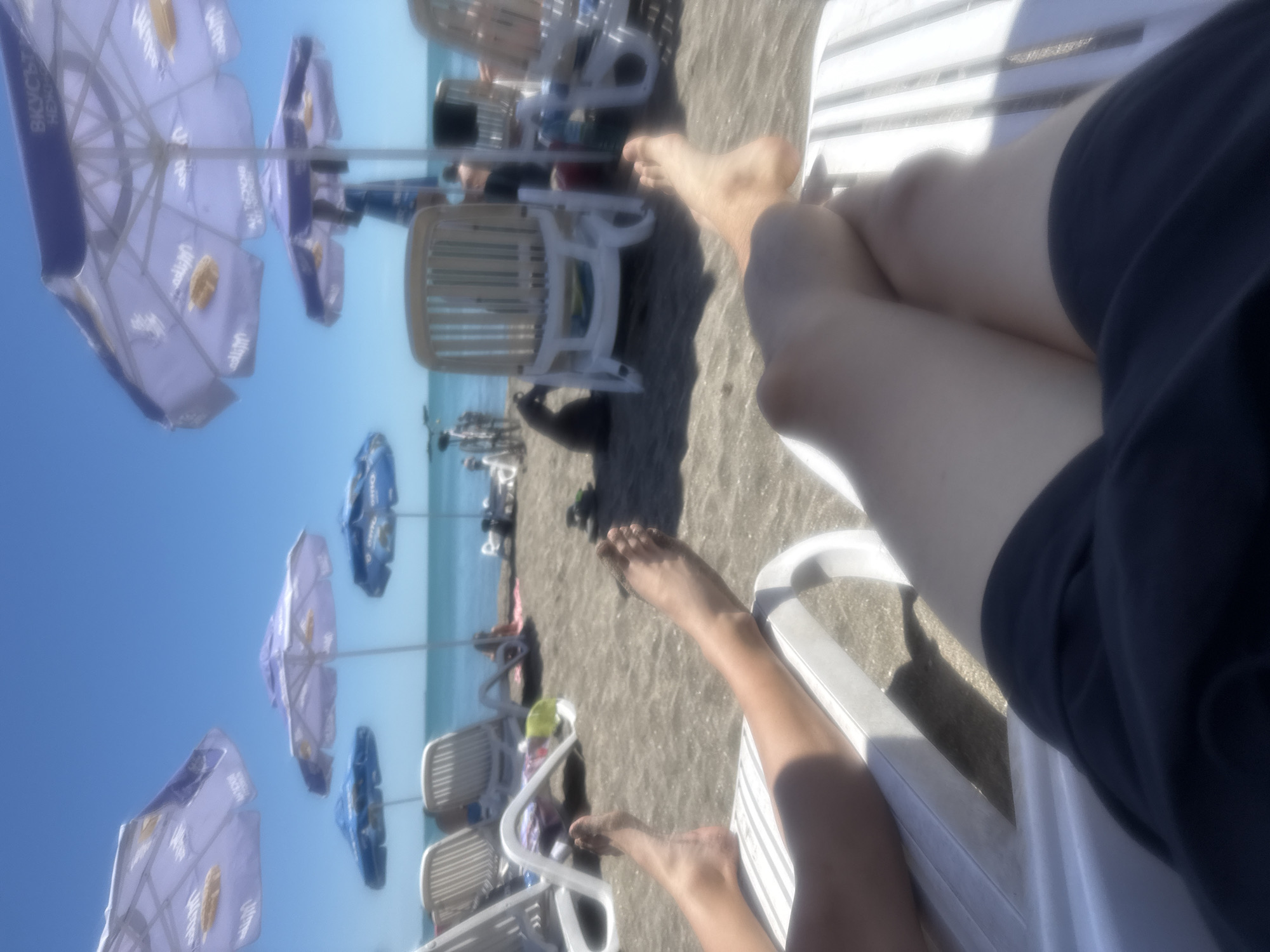 The image depicts a person relaxing on a white plastic lounge chair on a beach. The person is wearing dark blue shorts, and their legs are visible, crossed at the knees. The beach appears sandy, with multiple other lounge chairs and beach umbrellas in the background. The umbrellas are branded with a logo, possibly for a beverage or company. The sea is visible in the distance. The image was likely taken in Burgas, Bulgaria, known for its beaches.