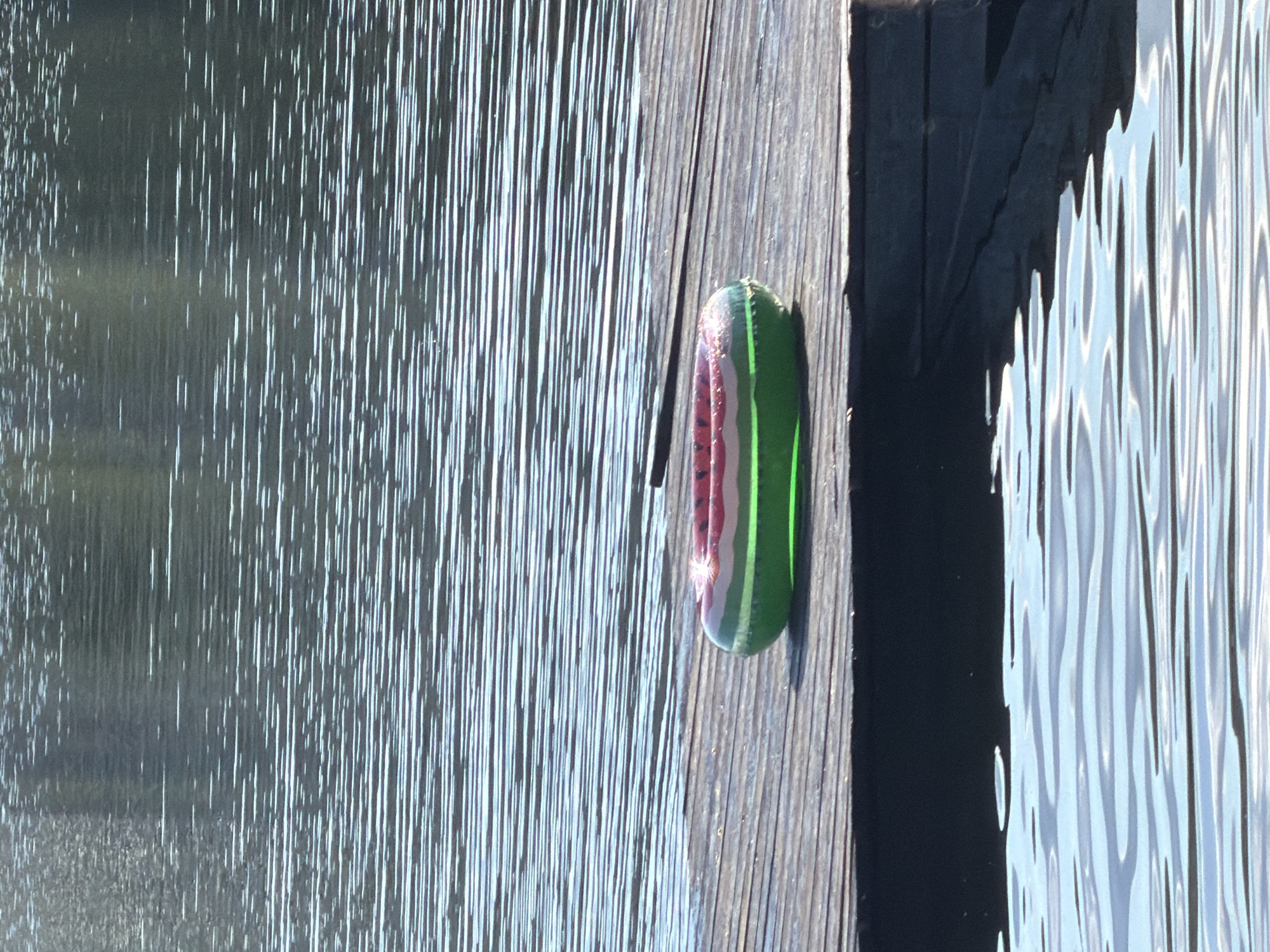 The image shows a close-up, high-angle shot of a wooden dock next to shimmering water. The dock planks are weathered and run vertically. A small, oval object with green and red stripes, resembling a miniature watermelon or a pool float, rests on the dock. The water is a light blue and appears rippled, with sunlight reflecting off its surface, creating vertical streaks of light. A dark shadow is cast from the dock into the water. The scene appears to be outdoors during the day, with bright sunlight. There is no visible text in the image.