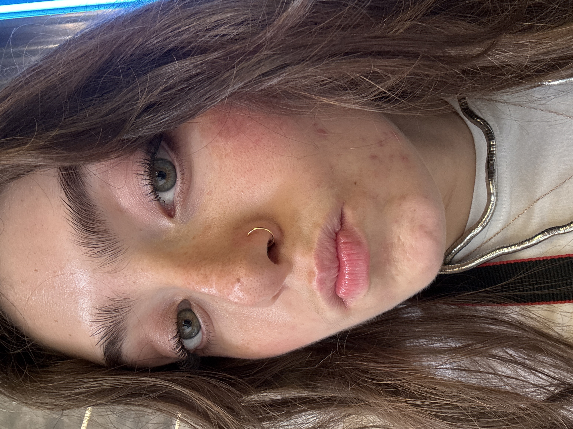 A close-up, slightly angled portrait of a person with light skin, brown hair, and green eyes. The person has a gold ring in their left nostril. Their hair is styled with loose waves, and the roots appear darker. The person is wearing a white top with a silver and black trim visible around the neckline. Skin imperfections such as freckles and minor blemishes are present. The background is out of focus but appears to be a light-colored surface, and a blue bar is visible at the top of the image. The location in the city of Barcelona, Spain, is not discernible from this image alone.