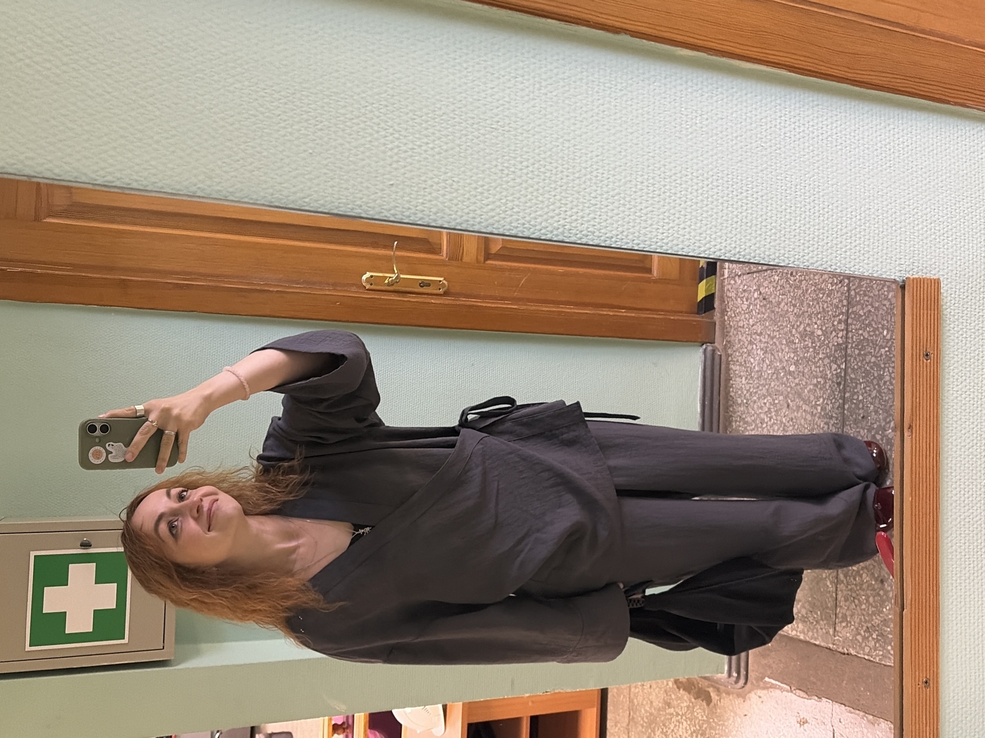 The image features a full-length mirror selfie of a woman indoors. She stands before the mirror, holding a light green smartphone to capture her reflection, and smiles subtly as she looks up. Her wavy, light brown hair frames her face.
She is dressed in a stylish, dark charcoal-grey two-piece set, which includes a relaxed-fit jacket or top with wide sleeves and matching wide-leg trousers. On her feet, she wears dark red or burgundy shoes. Her right hand, holding the phone, displays several rings on her fingers and a pink bracelet on her wrist. A delicate silver necklace is visible around her neck. The phone case has several small stickers, though their details are indistinct.
The background, reflected in the mirror, shows an indoor environment. A pale light-green wall with a textured pattern fills much of the upper and left parts of the reflection. A medium-brown wooden door, complete with panels and a brass coat hook, is visible behind her upper body. Below her left arm, a light grey first aid box with a prominent green cross symbol is mounted on the wall. The floor visible in the reflection is a light grey, speckled material, possibly terrazzo. A wooden frame borders the mirror on the right. The overall scene suggests a casual moment within a public or institutional building, such as a hallway or a changing area. No specific text is clearly legible, apart from the universally recognized green cross. This scene is located in Chaikovsky, Russia.