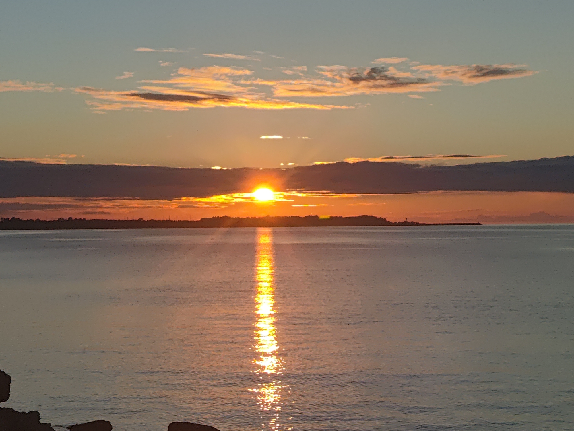 The image captures a serene sunset over the Baltic Sea, likely viewed from a coastal area in or near Tallinn, Estonia. The sun, a vibrant orb of golden light, is positioned just above a dark, horizontal band of clouds that stretches across the horizon. Rays of sunlight pierce through the clouds, illuminating the sky with hues of orange, yellow, and soft blue. The water in the foreground reflects the warm glow of the sunset, creating a shimmering path of light that stretches towards the viewer. A distant landmass, possibly the coastline of Tallinn, is visible as a dark silhouette against the colorful sky, with a faint outline of trees and structures. The overall atmosphere is one of tranquility and natural beauty.