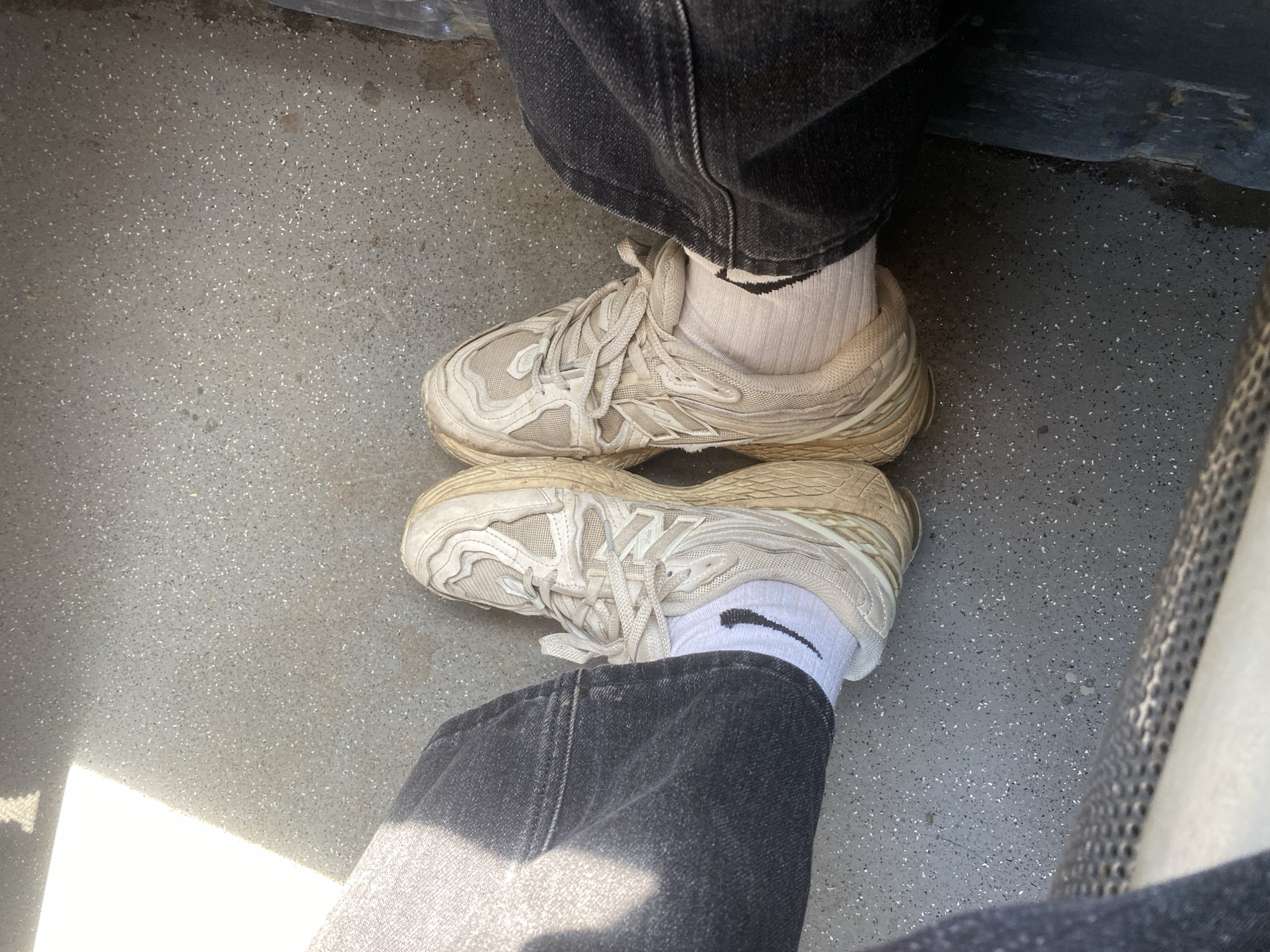 The image shows a person sitting, with their feet resting on the floor. They are wearing dark denim jeans and off-white athletic shoes with white socks that have a black Nike swoosh. The floor appears to be gray and speckled, possibly a public transportation setting like a bus or train. There is no information to support the location in Matyashivka, Ukraine.