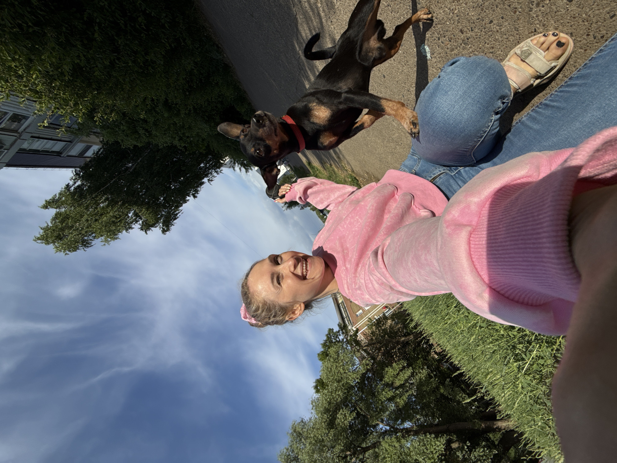 A woman in a pink sweater and blue jeans is walking a small, black and brown dog with a red collar on a leash. They are on a paved path, possibly a sidewalk or road, in a green, outdoor environment. Trees and a building are visible in the background. The sky is light blue with scattered white clouds. The location is in Chaikovsky, Russia.