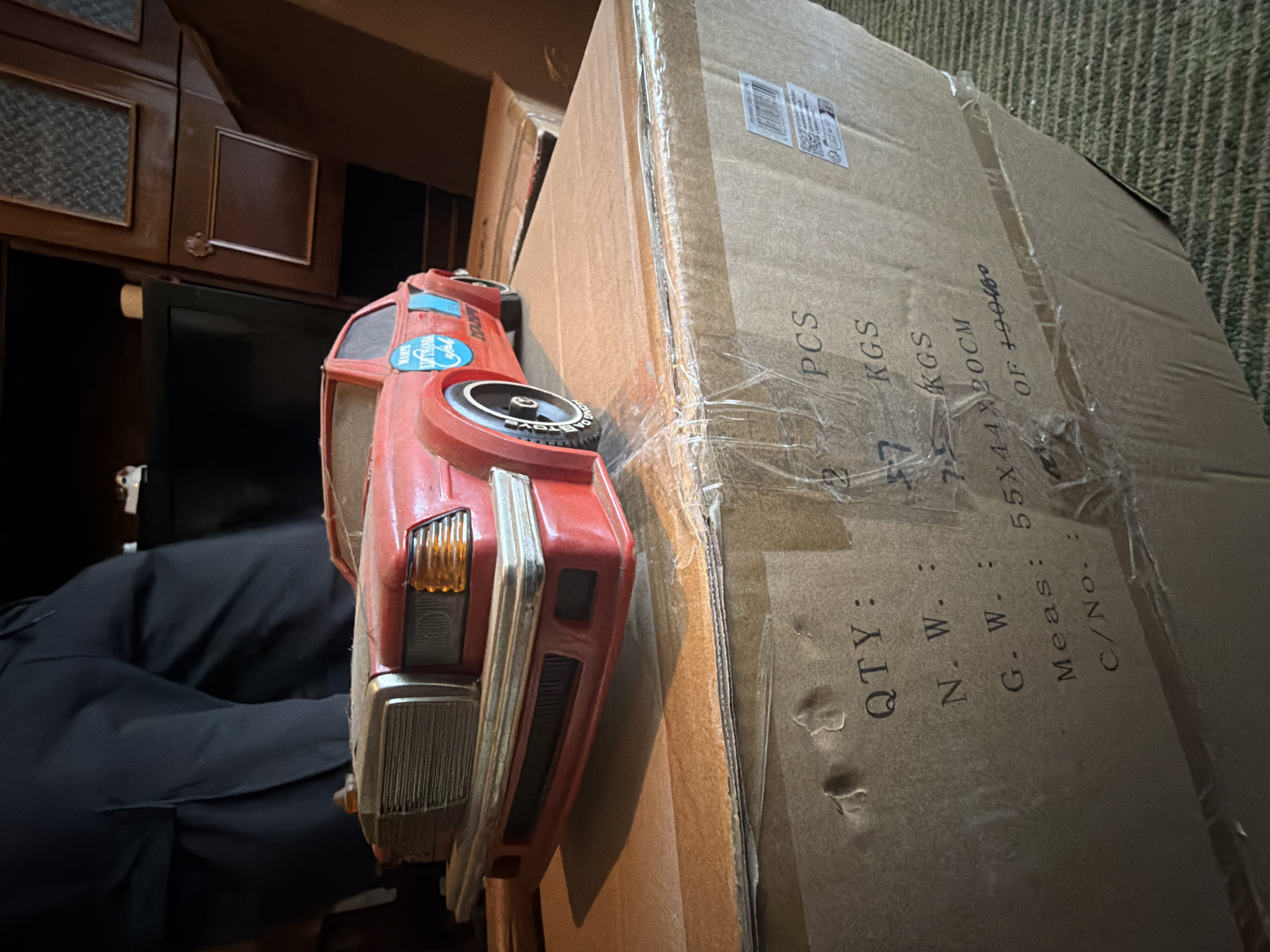 Here is a description of the image:
**Scene:** An indoor shot showing a cluttered area with various objects.
**Objects:** A large cardboard box, sealed with clear packing tape. The box has printed details including "QTY: 2 PCS", "N.W.: 7 KGS", "G.W.: 7 KGS", "Meas: 55X44X20CM", and "C/No.: OF 19040wo". Alongside the box is a red toy car, with a circular emblem in the center of one of its wheels. The background includes dark wooden furniture. Someone wearing black pants is partially visible.
**Environment:** The setting is likely an indoor storage area or a room with furniture. There's a carpet or rug visible in the background.
**Location:** Cannot be determined with the provided information. Sofia, Bulgaria cannot be confirmed from the image.