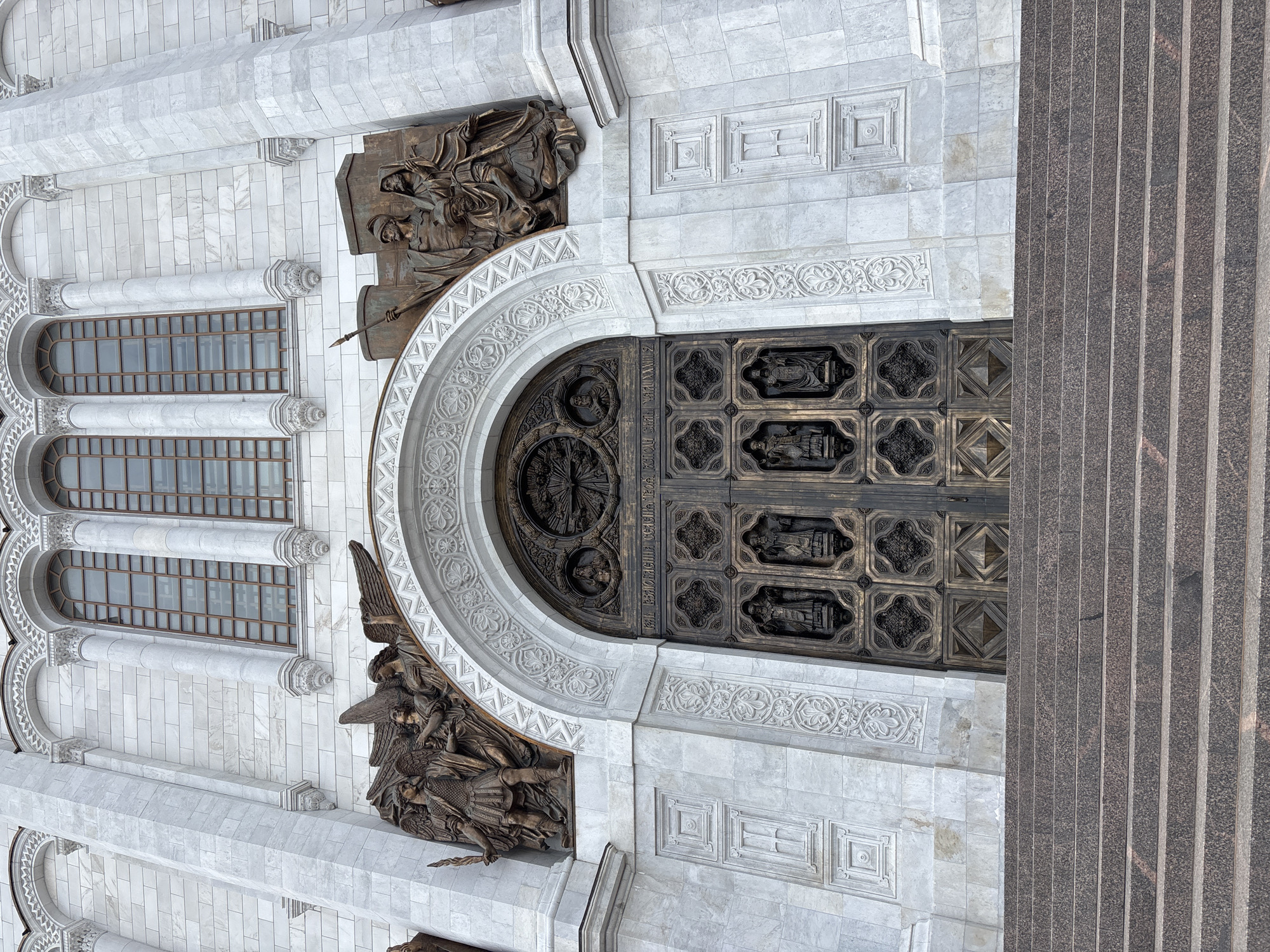 The image captures a detailed view of the Cathedral of Christ the Saviour in Moscow, Russia. The scene focuses on an ornate door and surrounding architectural elements. The door itself is dark and elaborately decorated with sculpted figures and geometric patterns. Surrounding the door, the building facade features a combination of light-colored stone or marble, sculpted friezes, and decorative moldings. Above the door, there is a bronze relief depicting a scene with multiple figures. Below the door, another bronze relief is visible featuring more figures. To the left of the door, there are arched windows with visible frames. A portion of a dark, granite pillar or facade element is visible on the right side of the image, adding to the grandeur and complexity of the building's design. The entire scene is captured in daylight.