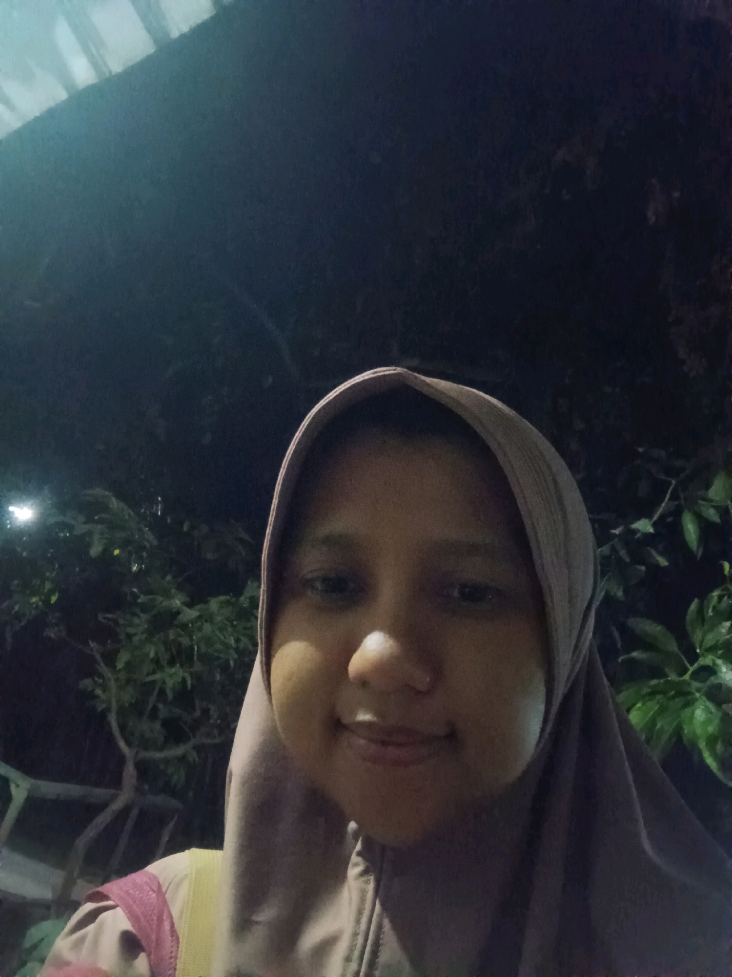 The image is a selfie taken at night, likely outdoors, with a person wearing a hijab. The background consists of dark foliage and some indistinct structures, suggesting a possibly rural or park-like setting. The lighting is dim, with a light source visible on the left side of the frame, illuminating some leaves. There is no information in the image to suggest the location is in Canga'an, Indonesia.