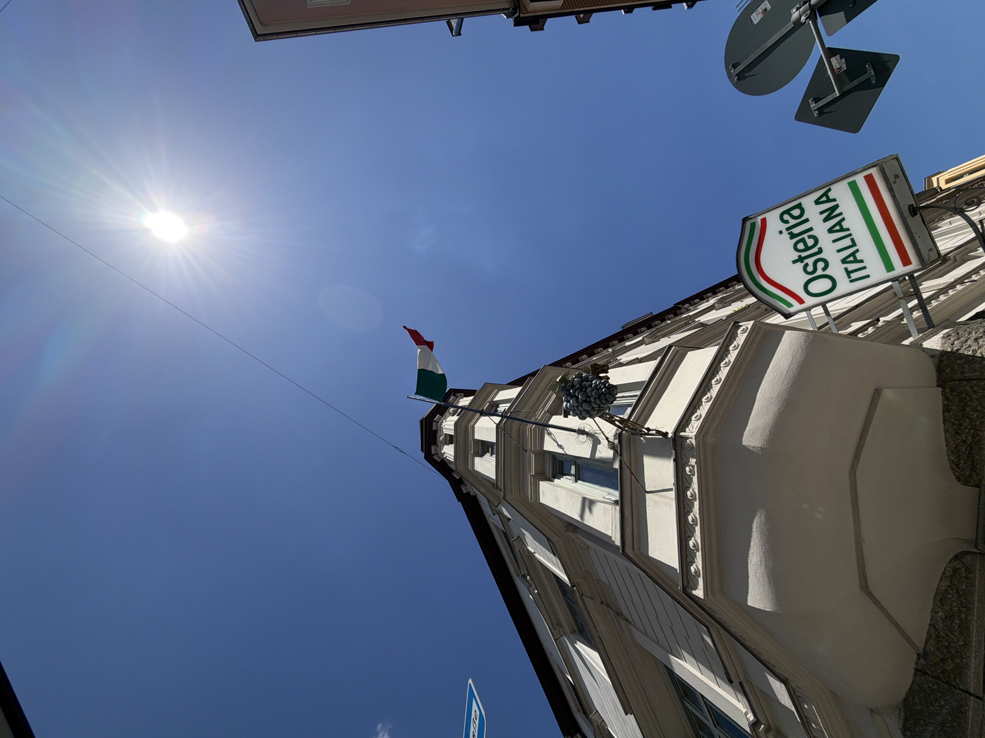 The image shows a view looking up towards a building in Munich, Germany, on a sunny day. The building is a light-colored, multi-story structure with architectural details. An Italian flag is visible, attached to a flagpole extending from the building. Below the flag, a decorative bunch of grapes is hanging. A sign with the text "Osteria ITALIANA" is also attached to the building, featuring the colors of the Italian flag. The sky is a clear blue with the sun shining brightly, creating lens flares. Other signs and parts of buildings are also visible in the background.