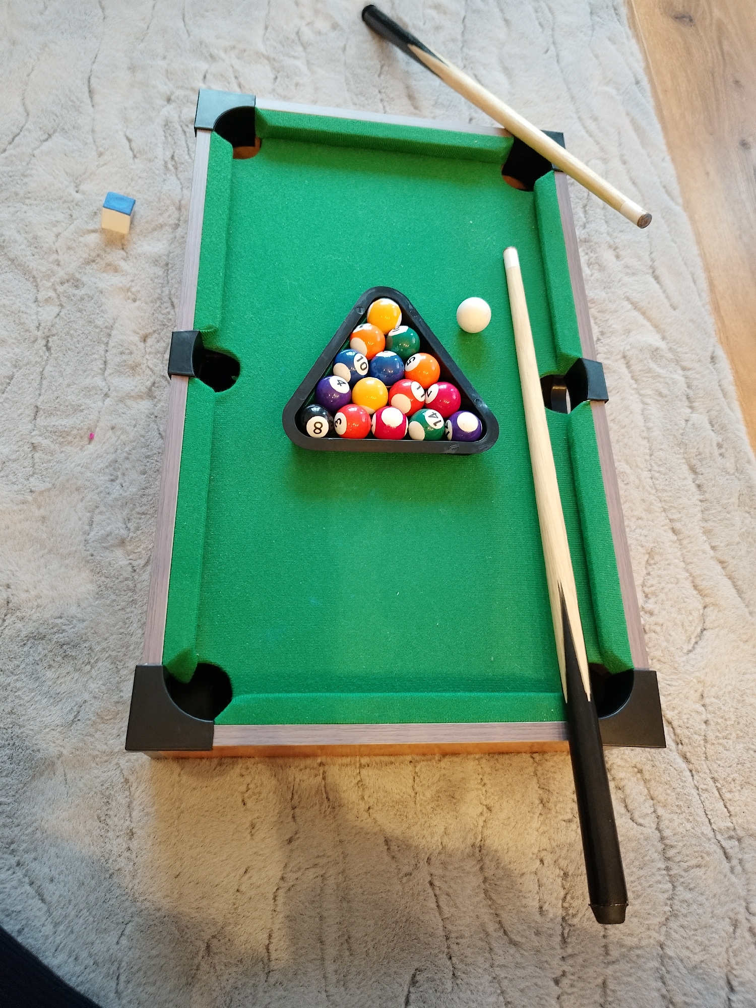 An overhead shot presents a miniature pool table positioned on a light-colored, shaggy carpet in an indoor setting. The table features a bright green felt playing surface, a light wooden frame, and black plastic pockets at each corner and along the long sides.
A complete set of billiard balls, including solids and stripes, is neatly racked in a black triangular frame, situated towards the center-right of the table. The white cue ball rests just outside the rack, to its left. Visible numbers on some of the balls include 8, 10, 14, and 4.
Two pool cues are laid out around the table. One cue extends diagonally across the upper left corner, partially on the table and partly off. The second cue lies along the right side of the table, parallel to the rail, with its black-gripped butt extending towards the bottom right. A small, light blue cube of pool chalk is placed on the carpet to the left of the table.
A section of a wooden floor is visible in the upper right background, adjacent to the carpet. The scene is brightly and evenly lit, suggesting an indoor environment. The arrangement of the equipment implies that a game of pool is either prepared to begin or has recently concluded. The location context is Sofia, Bulgaria.