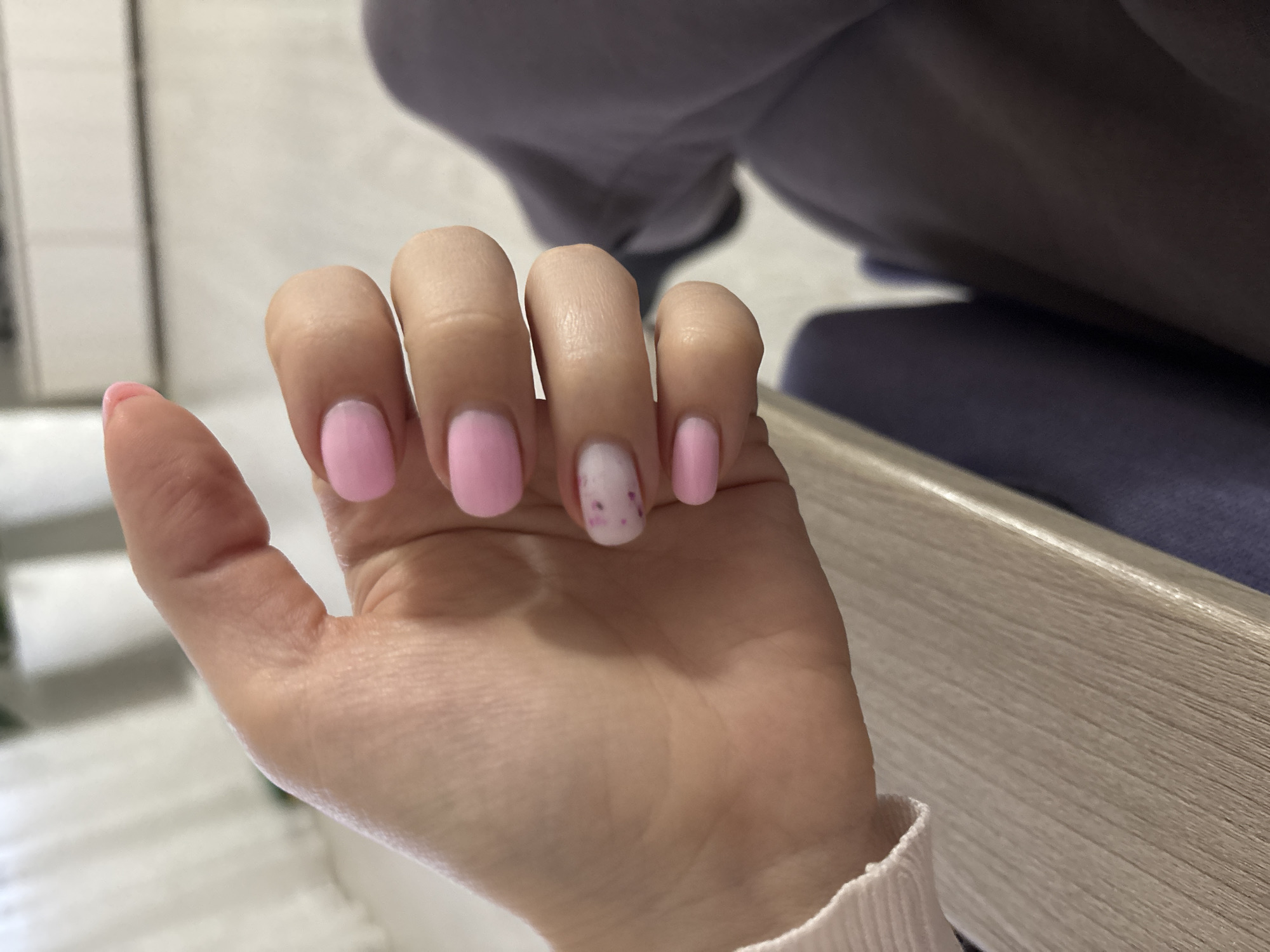 A close-up shot depicts a person's left hand, showcasing a manicure. The nails on the thumb, index, middle, and pinky finger are painted a solid light pink color. The nail on the ring finger is white with small, irregularly spaced pink dots. The hand is resting on a light-colored surface. The background is blurred and shows clothing (likely a gray sweater and a dark blue chair) and a white wall. The location is not discernible from the image. The location specified in the prompt is impossible to determine from the image.