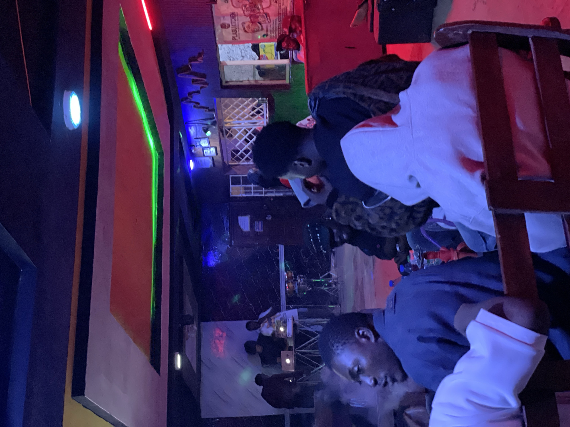 In Durumi, Nigeria, this image captures a dimly lit interior scene, likely a bar or entertainment venue, at night. Several people are present, some seated and others standing, with a focus on two individuals in the foreground. One man in a dark blue shirt is leaning forward, his face partially obscured, while another person in a white garment is seated in a wooden chair. The ambiance is enhanced by colorful neon lights, including vibrant green and red, casting an atmospheric glow on the surroundings.
In the background, a small stage area is visible with musical instruments and lighting equipment, suggesting a live performance or DJ set is underway. There are also shelves with items that could be drinks or food items, hinting at a place that serves refreshments. The overall mood appears relaxed and social, typical of a night out at a local establishment. The presence of multiple individuals and the entertainment setup indicate a gathering or event taking place. The lighting, predominantly artificial and colorful, suggests the event is occurring indoors after dark.