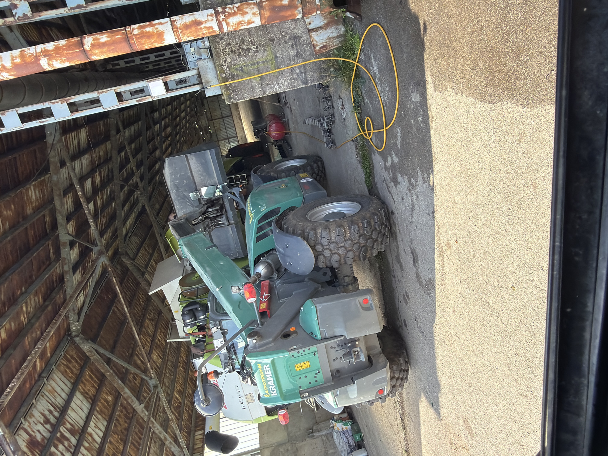 This image, captured in Bazan, Bulgaria, depicts an industrial or agricultural setting, likely inside or at the entrance of a large workshop or shed. The main subject is a green and grey Kramer telehandler or compact loader, prominently parked on a concrete surface. The vehicle features large, rugged tires and has "KRAMER" clearly visible on its side in white lettering, with smaller text above it appearing to read "APOTHEUM" and a yellow warning symbol. Another piece of green machinery, possibly a tractor or additional equipment, is partially visible behind the Kramer, bearing the text "P 12" and what looks like part of "CLAAS".
Overhead, a network of aged, rusted metal pipes and grey support structures extends across the ceiling, suggesting a processing or storage facility. A bright yellow hose is coiled on the ground, extending from a red air compressor positioned behind the Kramer vehicle. The scene is illuminated by strong natural daylight, indicating it is daytime, which casts distinct shadows on the concrete floor. The ground itself is a mix of concrete and what appears to be a coarser, lighter-colored aggregate. No individuals are present in the image.