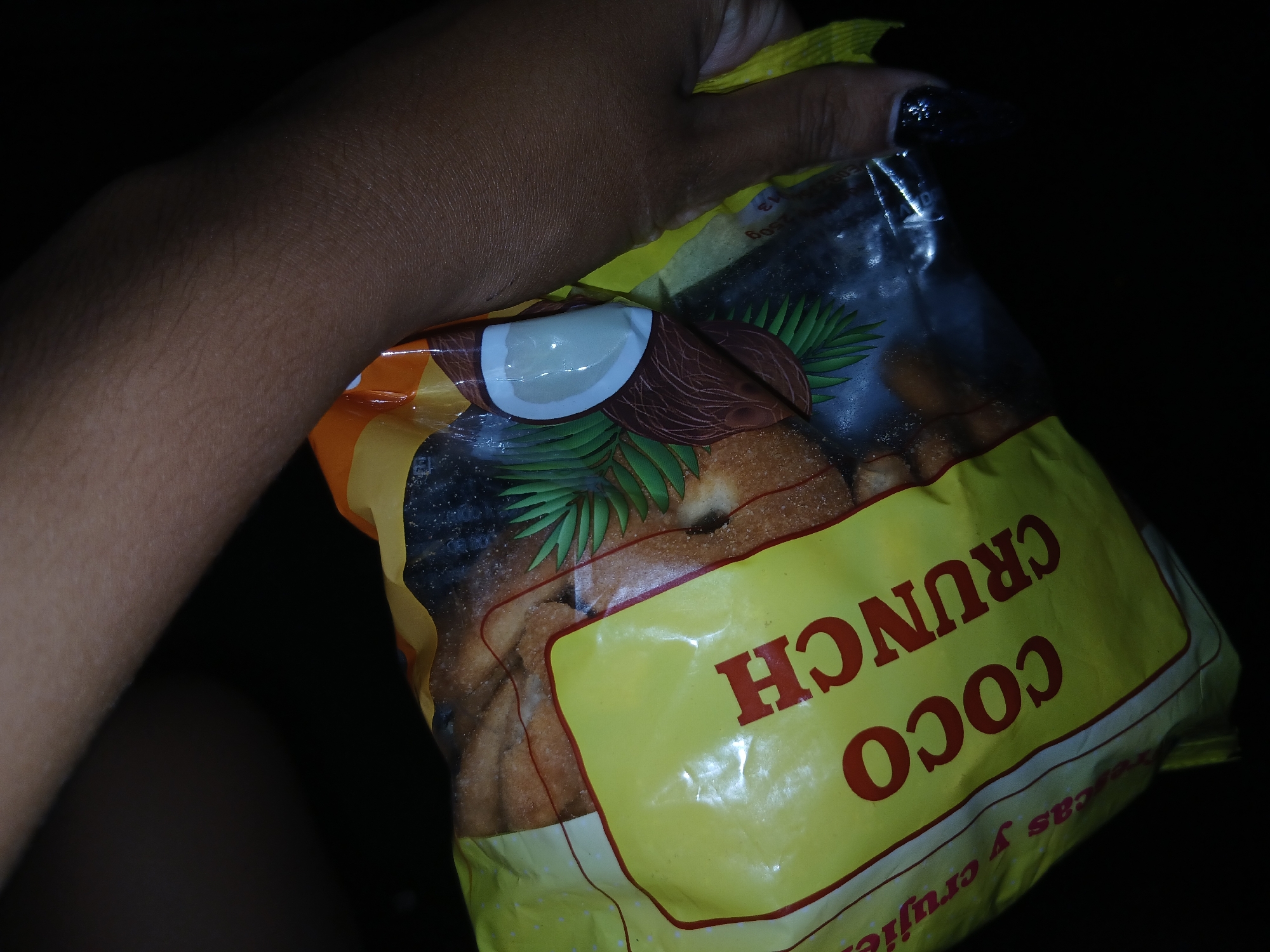 A person is holding a bag of "Coco Crunch" cookies in a dark setting. The bag features a picture of a coconut and coconut leaves. The person has dark skin and dark nail polish. The text on the bag reads "COCO CRUNCH". The location of the image is unknown, and no information is available to infer it's in Altagracia, Venezuela.