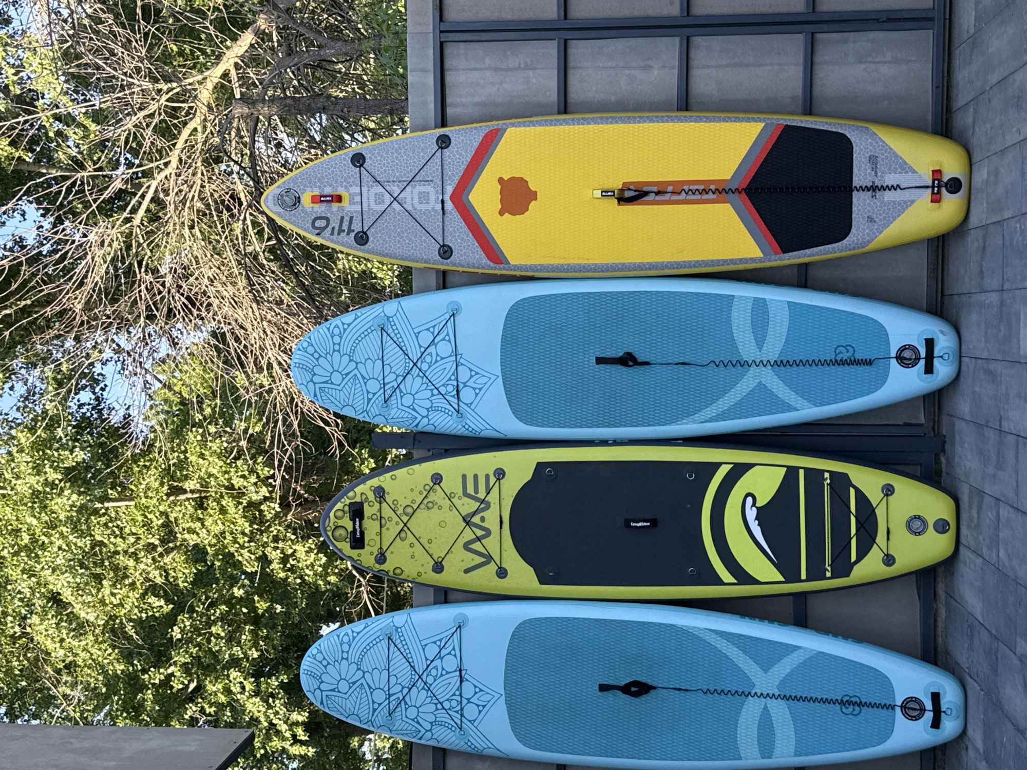 Four paddleboards are stacked vertically on a gray rack against a gray, stone-textured wall. The top board is yellow with gray, red, and black accents. The second and fourth boards are light blue with mandala-like designs on the front. The third board is yellow-green with black accents and the word "WAVE" in white and black. Trees and sky are visible in the background. The location is not identifiable as Rostov-on-Don, Russia, based on the provided image.