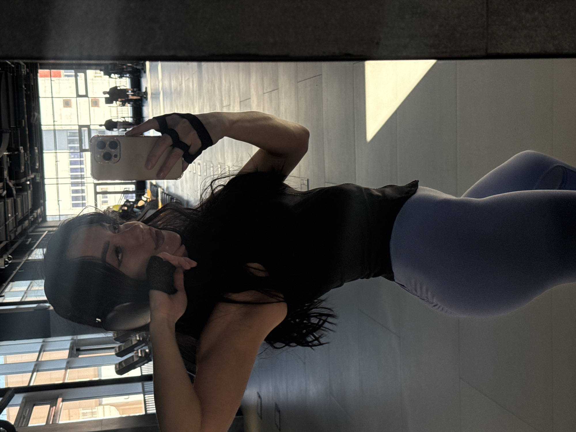 The image is a mirrored selfie of a woman in a gym. She is wearing a black top, blue leggings, and has headphones around her neck. She is holding a smartphone in her right hand, using it to take the photo. The mirror reflects the gym environment, which includes workout equipment and windows showing a cityscape. Outside the windows are several tall buildings and blue skies. There is a large sunbeam reflected in the mirror on the right side of the picture. The location is indicated to be Krasnoyarsk, Russia, based on visual elements such as the urban architecture visible through the windows.
