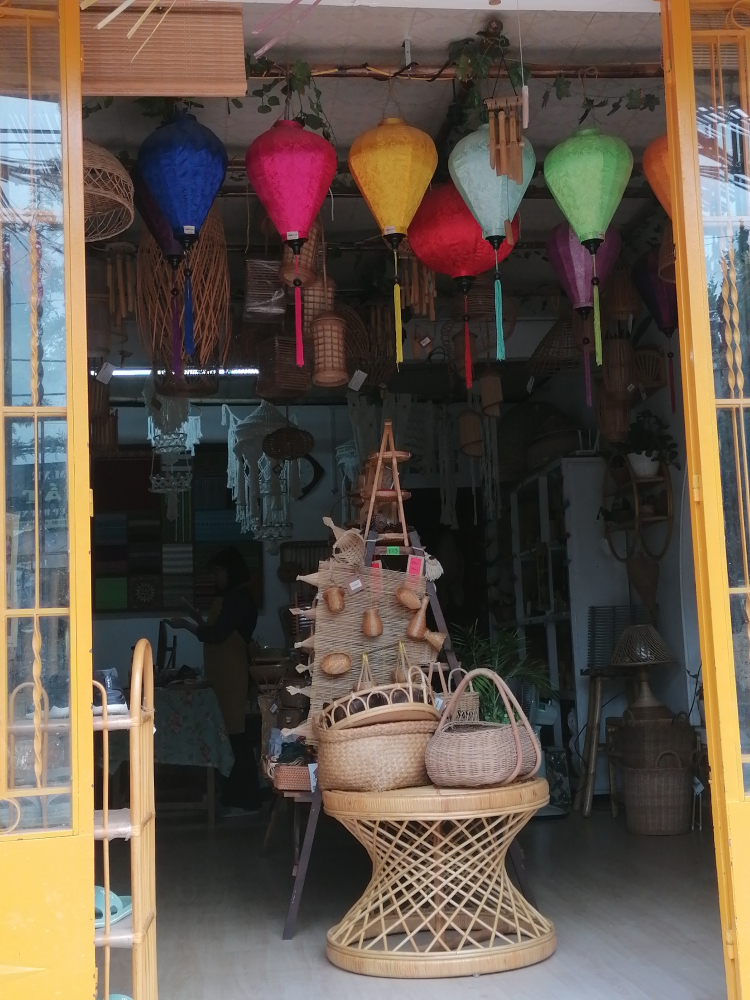The image depicts the entrance to a shop in Lang Biang - Đà Lạt, Vietnam, specializing in handcrafted bamboo and rattan products. The shop's exterior features a bright yellow frame for a door and window, providing a vibrant contrast to the goods displayed within. Numerous colorful paper lanterns in various shapes and sizes hang from the ceiling, including blue, pink, yellow, light blue, and green. These lanterns are interspersed with an array of woven baskets and other decorative items, also made from natural materials like bamboo and rattan.
Inside the shop, a person is partially visible in the background, appearing to be a shopkeeper or employee. They are wearing dark clothing and are standing near shelves filled with more products. The shelves themselves are constructed from wood or bamboo and are laden with a variety of items, including more baskets, textiles, and possibly wind chimes or other decorative pieces. A patterned tablecloth is visible on a table behind the person.
In the foreground, a large woven rattan table with a unique, hourglass-like design serves as a display platform. It holds two prominent woven baskets, one larger and rounder, and a smaller, more oval-shaped basket with handles. A tiered shelving unit made of light-colored wood or bamboo stands to the left of the entrance, displaying smaller woven items and possibly some footwear or personal accessories on its shelves. The floor appears to be a smooth, light-colored surface. The overall impression is one of a well-stocked artisanal shop offering a variety of decorative and functional items made from natural materials.