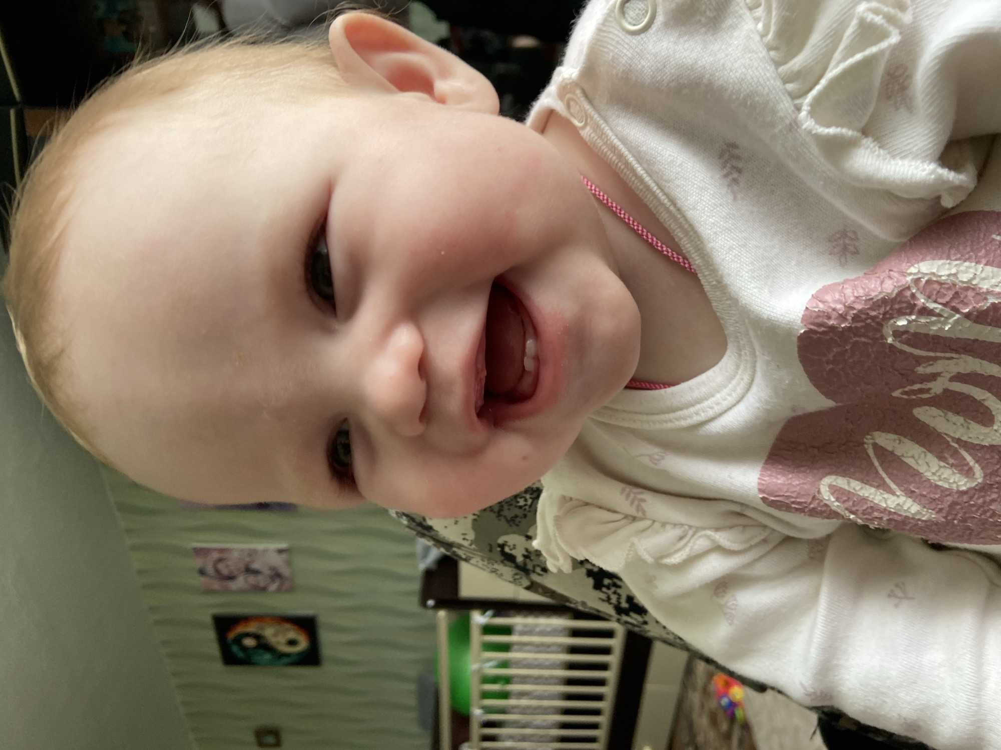 A baby is shown up-close, turned slightly to the side, and smiling with an open mouth. The baby has fair skin and blonde hair. The baby is wearing a light-colored shirt with pink details. The background appears to be a room with decorations on the wall. The location mentioned, Фоки, Russia, is not directly visible in the image, and therefore cannot be confirmed.