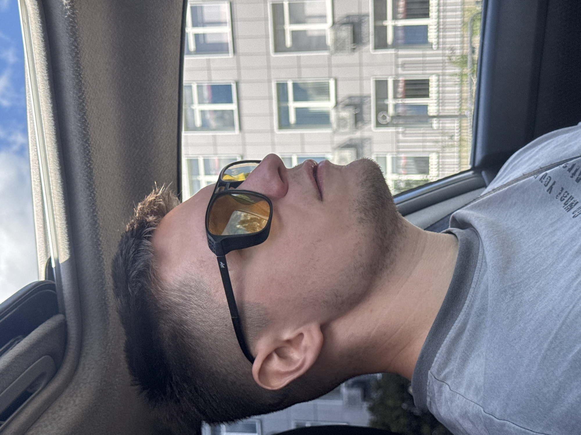 A man with short, dark hair and light skin is in the passenger seat of a car, looking to the right. He is wearing dark-rimmed sunglasses with yellow lenses and a light grey t-shirt. The car's sunroof is open, revealing a blue sky with white clouds. In the background, a building with multiple windows and balconies is visible. The scene appears to be in the city of Chaikovsky, Russia.