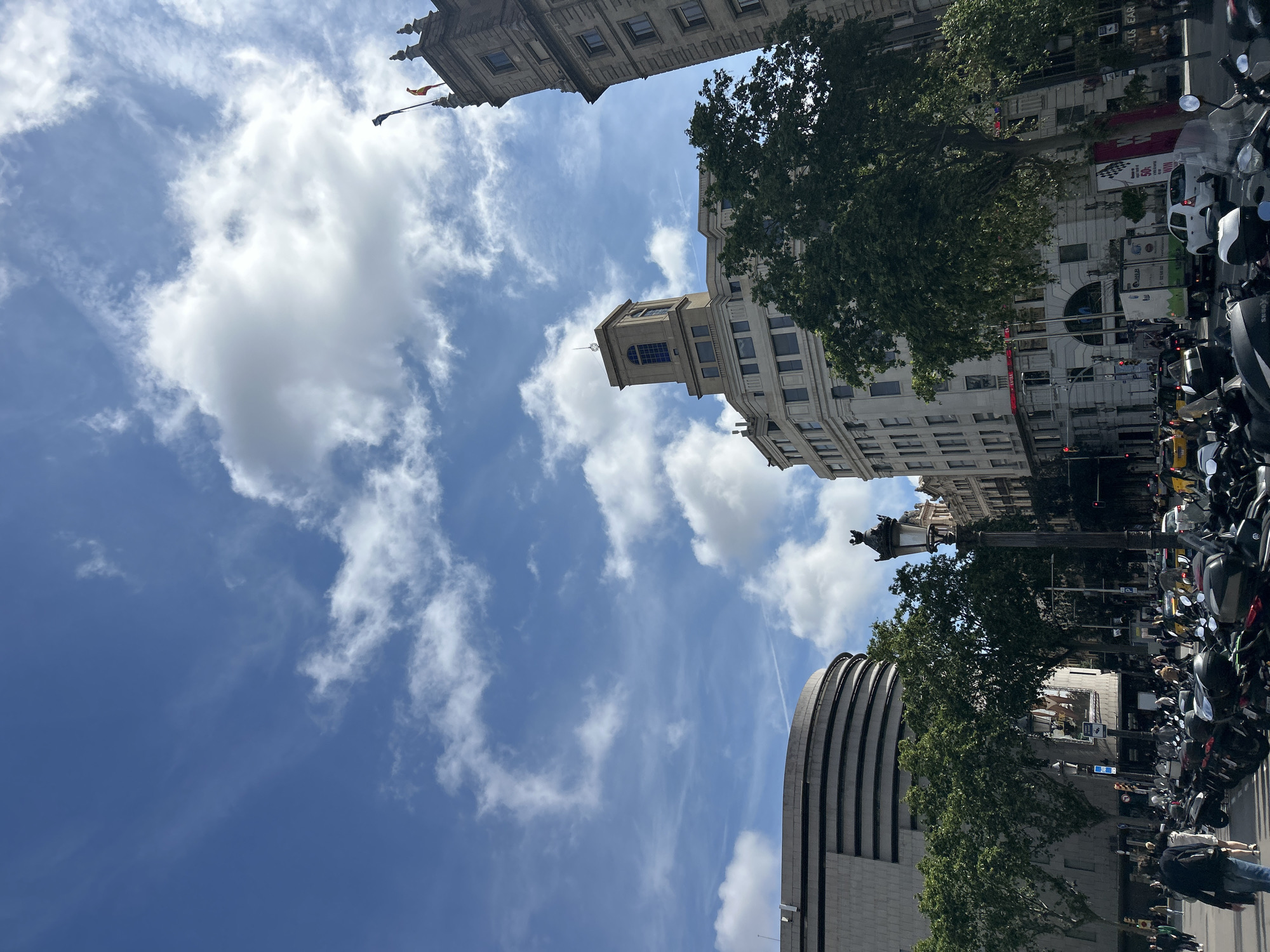 This image shows a street view in Barcelona, Spain, taken from a low angle looking upwards. The sky is blue with scattered white clouds. The surrounding architecture includes historical buildings with ornate detailing and modern, multi-story buildings. Trees with lush green leaves frame the scene, partly obscuring the buildings. On the right side of the image, a street lined with parked motorcycles and some pedestrians is visible. A tall monument with a statue on top rises into the frame. The scene is bathed in daylight, suggesting it was taken during the day.