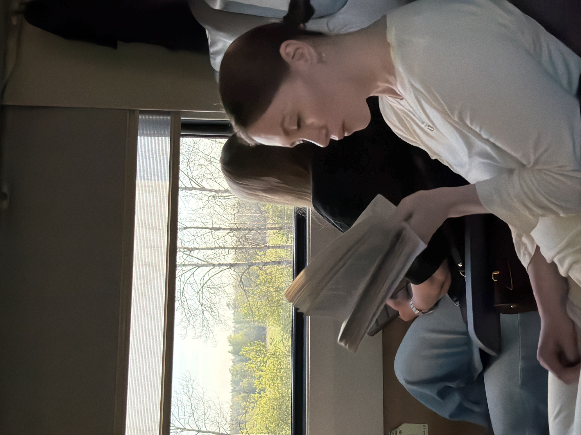 A photograph taken on a train in Ushaki, Russia, shows two individuals seated by a window. The person in the foreground has dark hair pulled back in a bun and is wearing a white top. They are reading a document. The second individual, seated next to them, has blond hair and is wearing a black shirt and blue jeans. A brown bag rests next to them. Through the window, a view of trees and a partially-obscured landscape are visible.