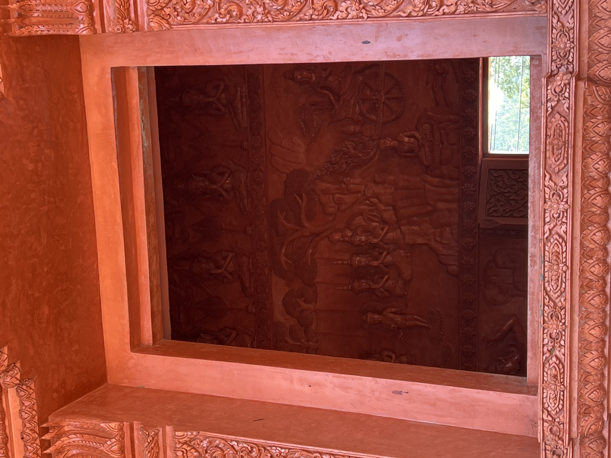 The image shows the exterior of a building with intricate orange-red ornamentation. A large rectangular opening reveals a relief carving on the interior wall. The relief depicts several figures in a narrative scene, including figures in prayer, a person on a chariot, and other individuals in stylized poses. The relief is also predominantly orange-red in color. Decorative carvings are visible around the opening and along the exterior facade. The overall impression is of a richly decorated temple or cultural building. The image's context suggests a location in Baan Hua Thanon, Thailand, known for its Buddhist temples and artistic traditions.