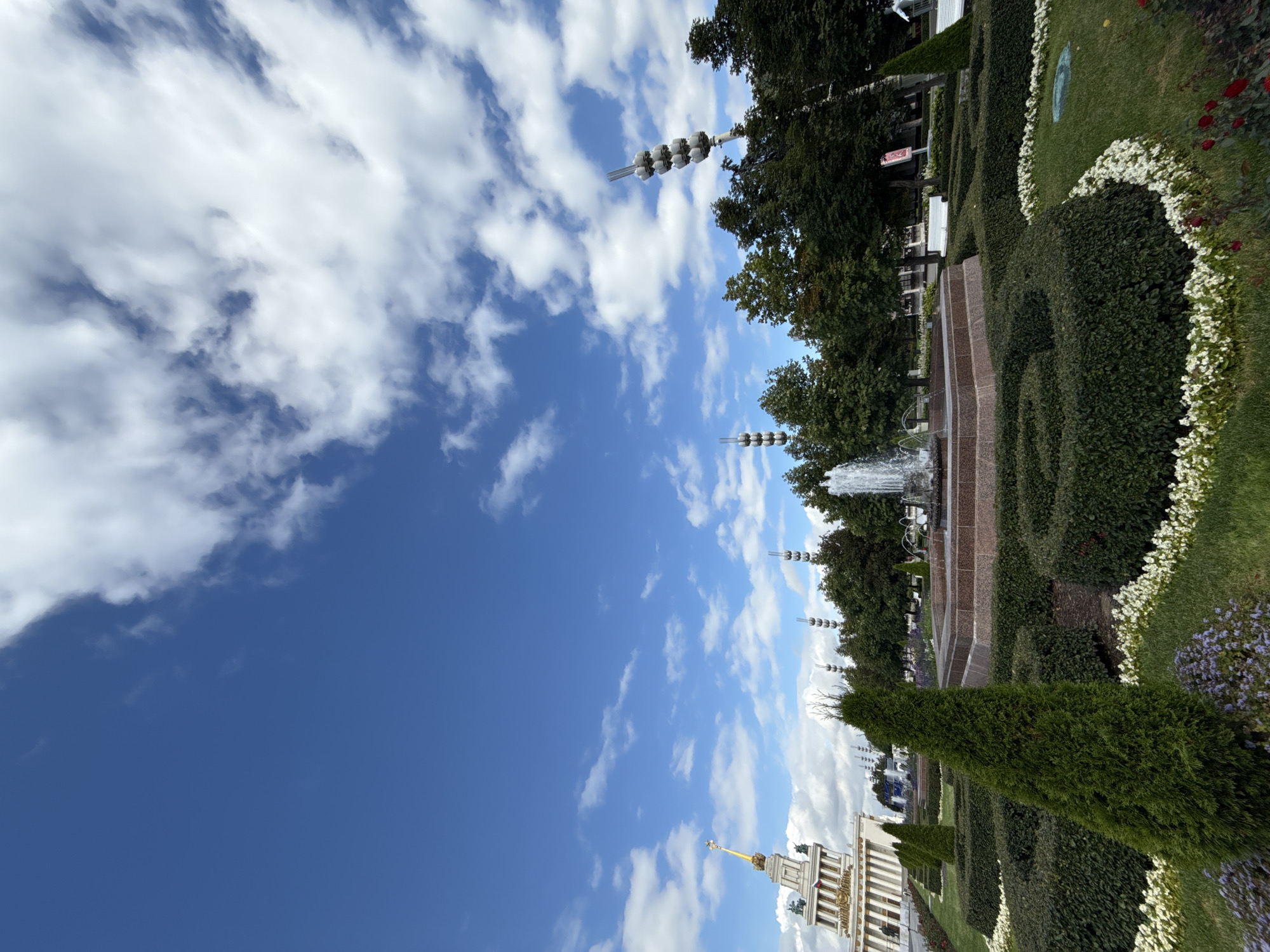 This image captures a view of the VDNKh park in Moscow, Russia, on a bright, cloudy day. In the foreground, a manicured garden features several tiered bushes, forming geometric shapes, with white and purple flowers lining the pathways. A large, octagonal fountain made of granite sits in the middle ground, with water spouting from its center. Behind the fountain, a dense line of green trees forms a backdrop, interspersed with distinctive tall, slender structures topped with clusters of lights. To the left, a grand, ornate building with a golden spire is visible, a prominent architectural landmark. The sky above is a vibrant blue, dotted with fluffy white clouds. The scene depicts a well-maintained public park, likely during daylight hours, with no people visible.