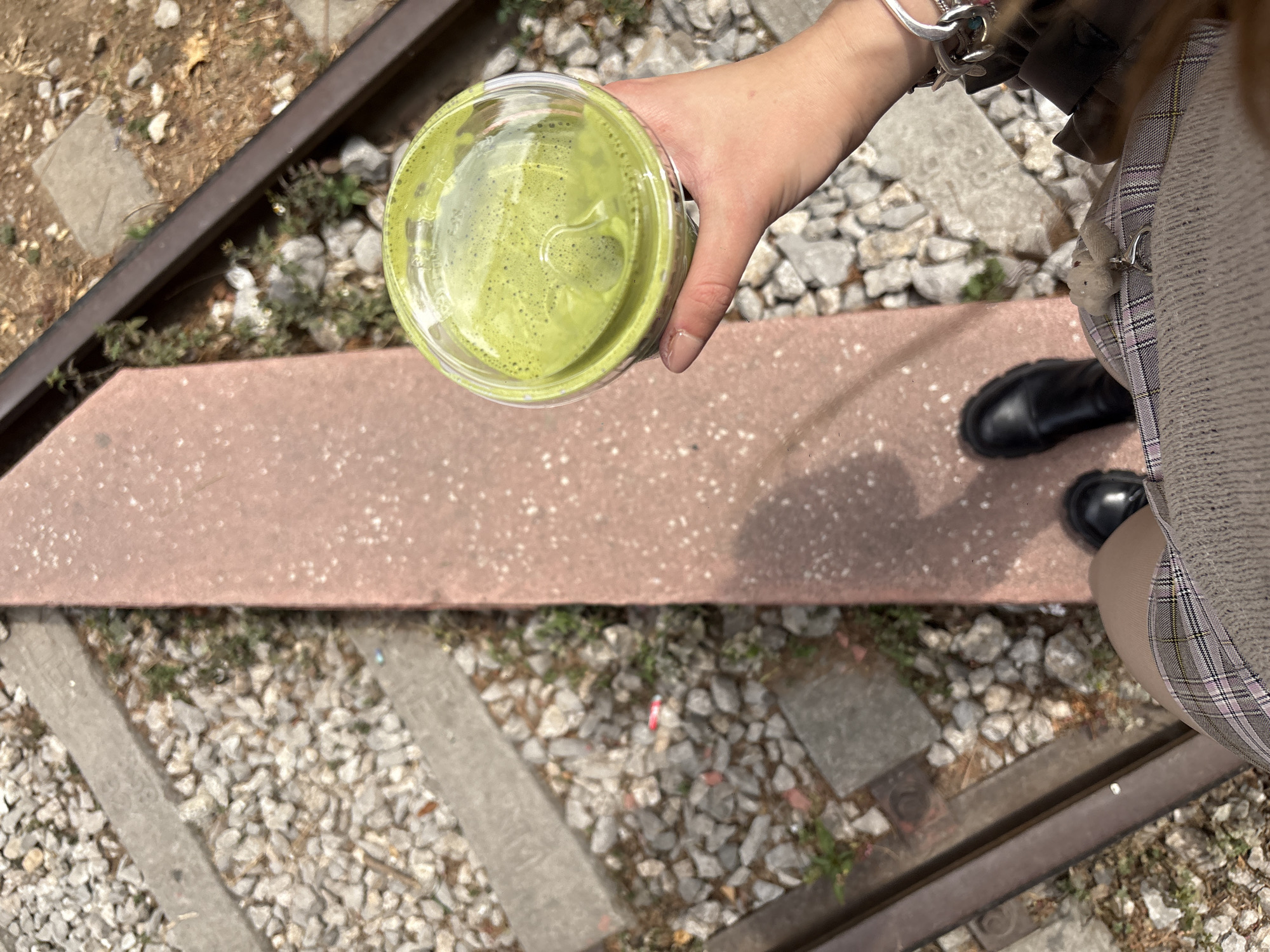 A high-angle view shows a woman standing on a disused railway track with gravel between the sleepers. She holds a disposable transparent plastic cup filled with a green liquid (possibly matcha) in her left hand. She is wearing a plaid skirt, tan tights, and black boots. Her left wrist has several silver bracelets. The scene is in daylight, suggesting an outdoor setting. Based on the presence of disused railway tracks, this may be a location in Mexico City known as the "Ciclovía Ferroviaria." The lighting conditions suggest a daytime setting.