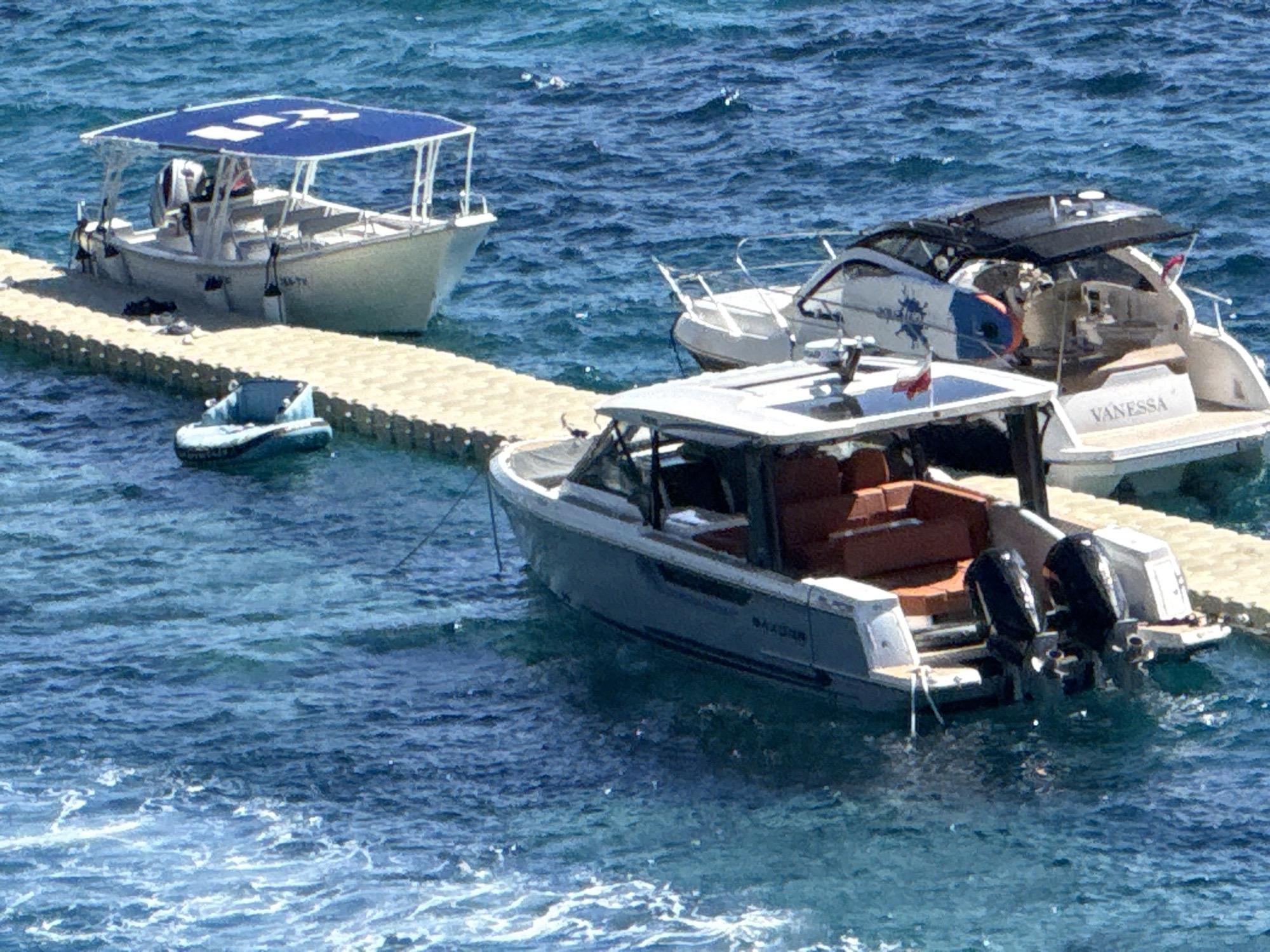 The image shows a dock with three boats on a bright, sunny day in Budva, Montenegro. A small, light blue inflatable dinghy is tied to a pontoon dock. Next to it, a white boat with a blue canopy is also docked. Further out in the water, a white yacht named "VANESSA" is anchored. In the foreground, a gray speedboat with twin outboard motors is positioned in the water, slightly to the right of center. The water is a clear, vibrant blue, with some whitecaps suggesting light waves. The scene appears to be a recreational or marine setting, likely a marina or a popular spot for boating.