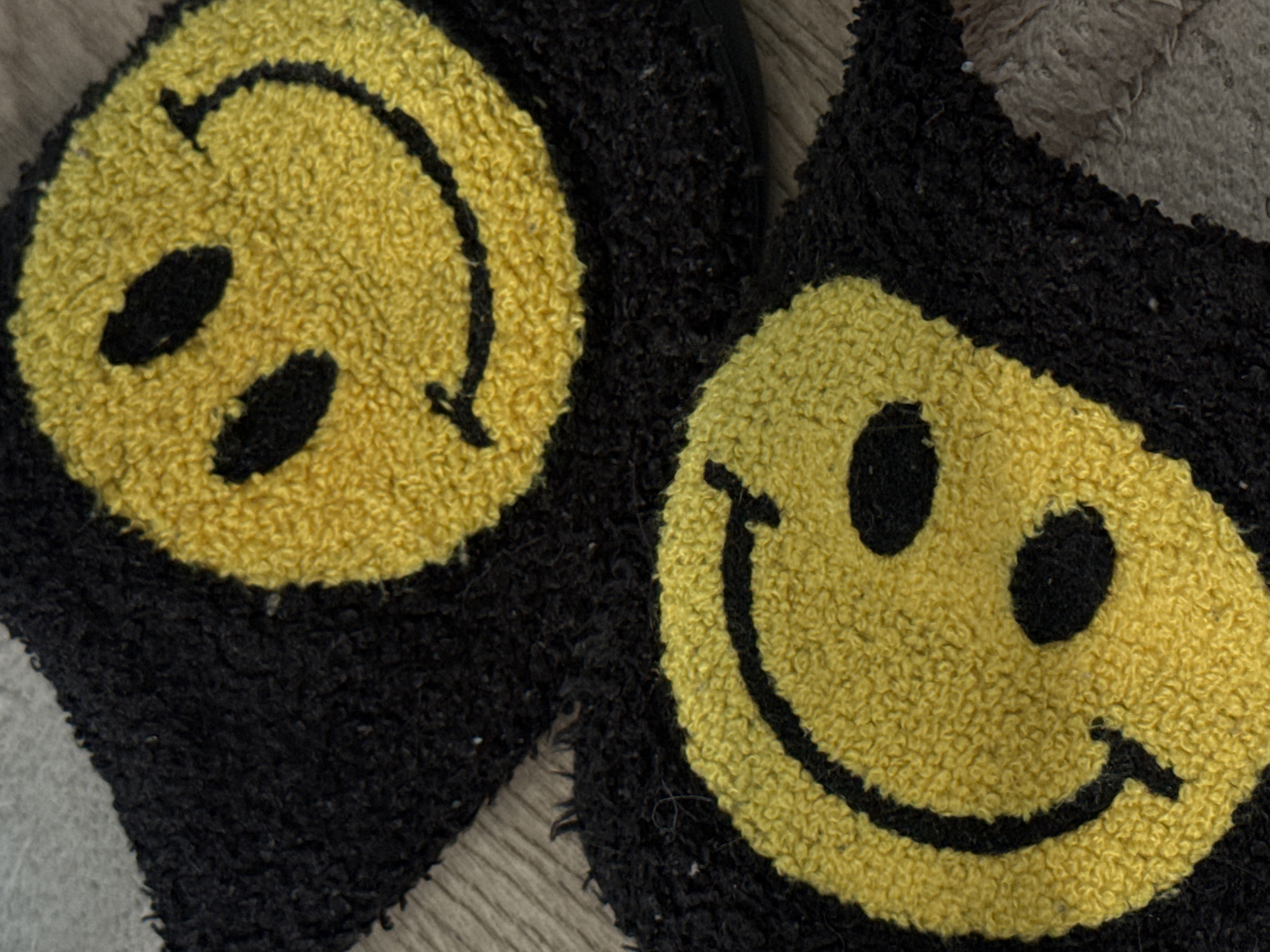 The image shows two black slippers with yellow smiley face designs. The faces are made of a fuzzy material and have black details for the eyes and mouth. The slippers are resting on a textured surface, likely indoors. There is no indication of the location being in Khimki, Russia.