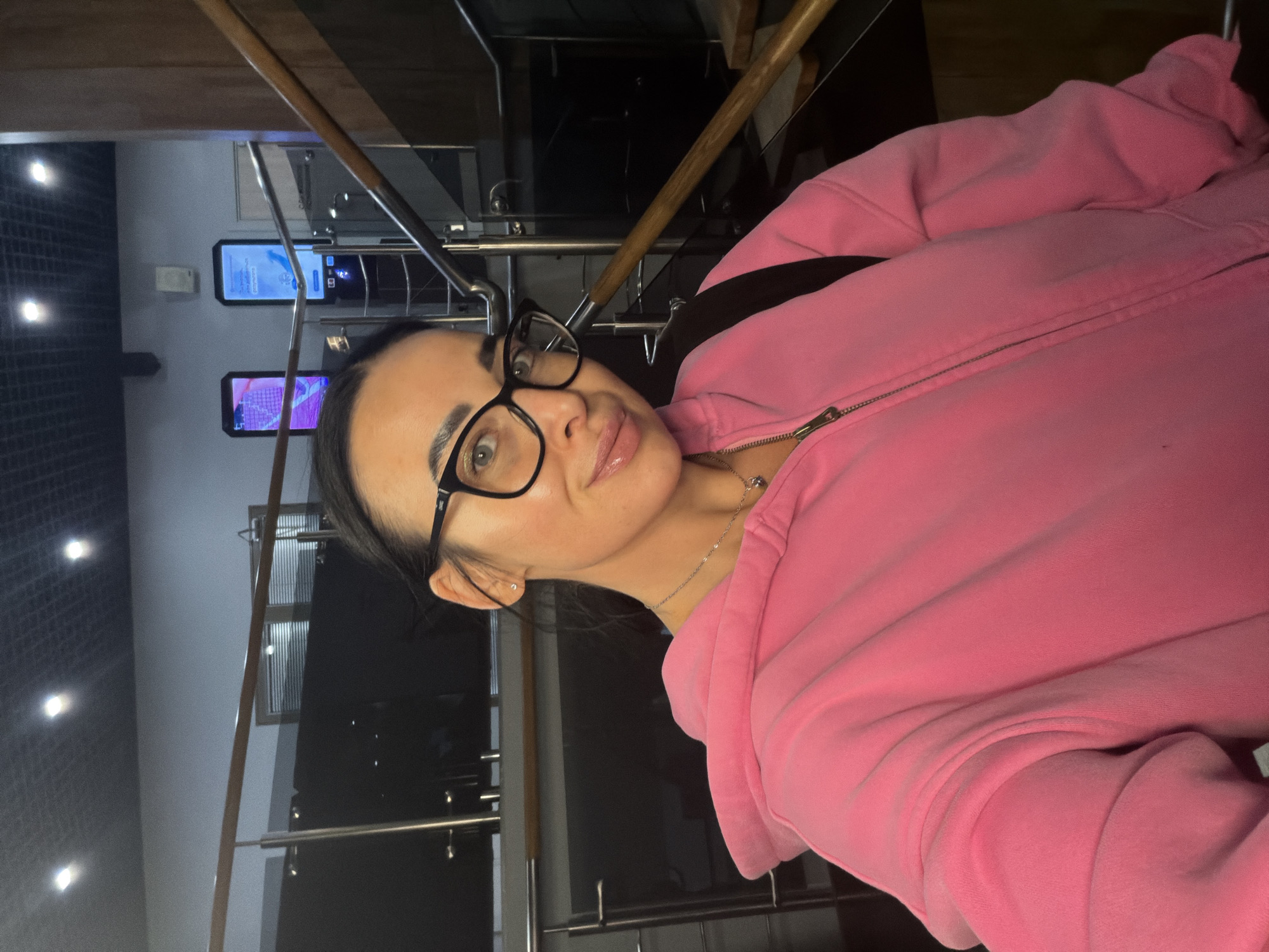 A woman with dark hair pulled back and wearing black glasses and a pink zip-up hoodie takes a selfie. She is standing indoors. Behind her is a railing system with metal and wood components. In the background, there are digital screens displaying information. The location is likely an indoor space in Saint Petersburg, Russia.