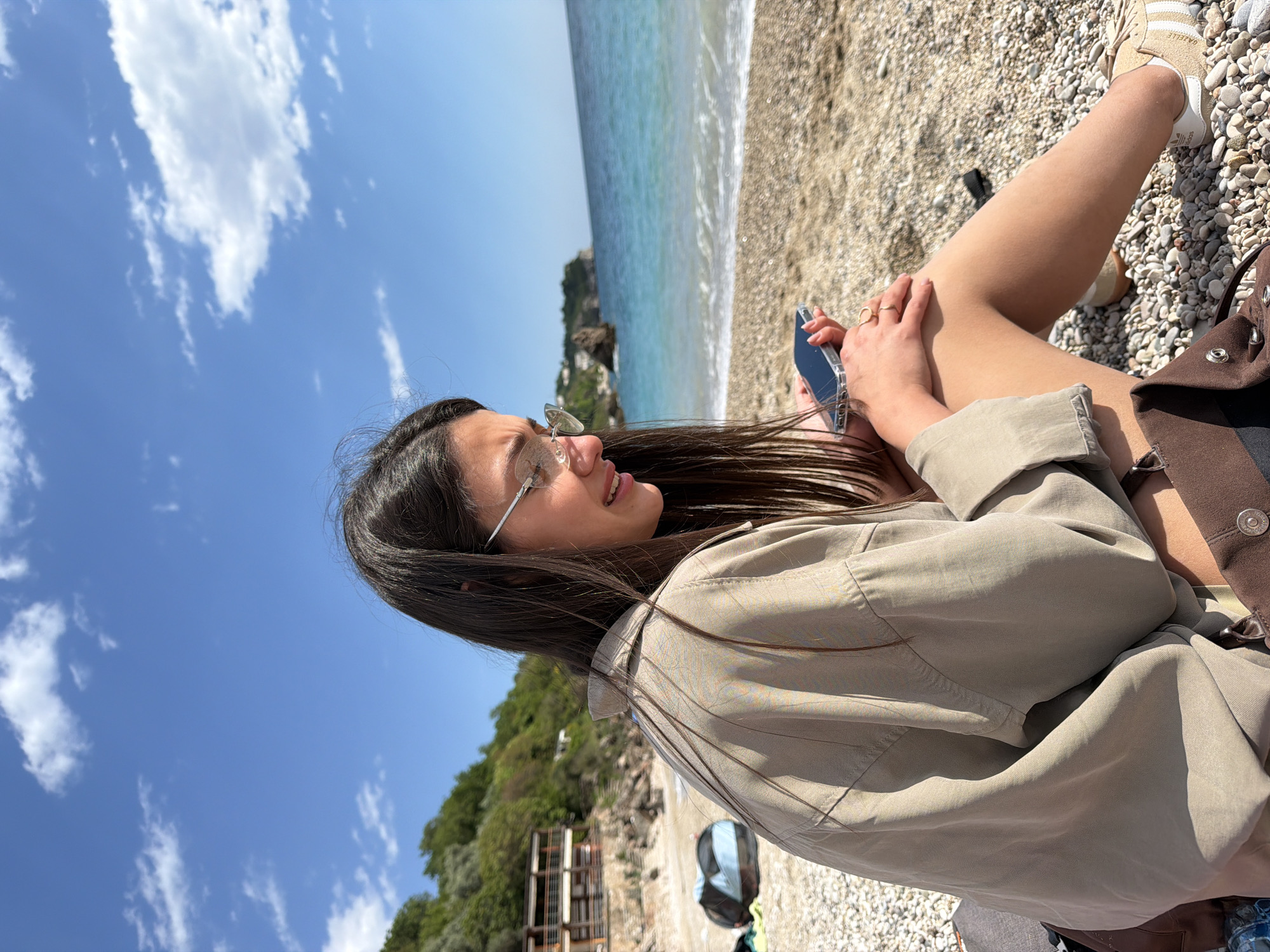 A young woman sits on a pebble beach in Drobnići, Montenegro, overlooking the Adriatic Sea under a partially cloudy blue sky. She wears sunglasses, a beige shirt, and Adidas Gazelle sneakers. She is holding a phone in her hand, and a brown bag is beside her. Lush green vegetation covers the nearby hills.