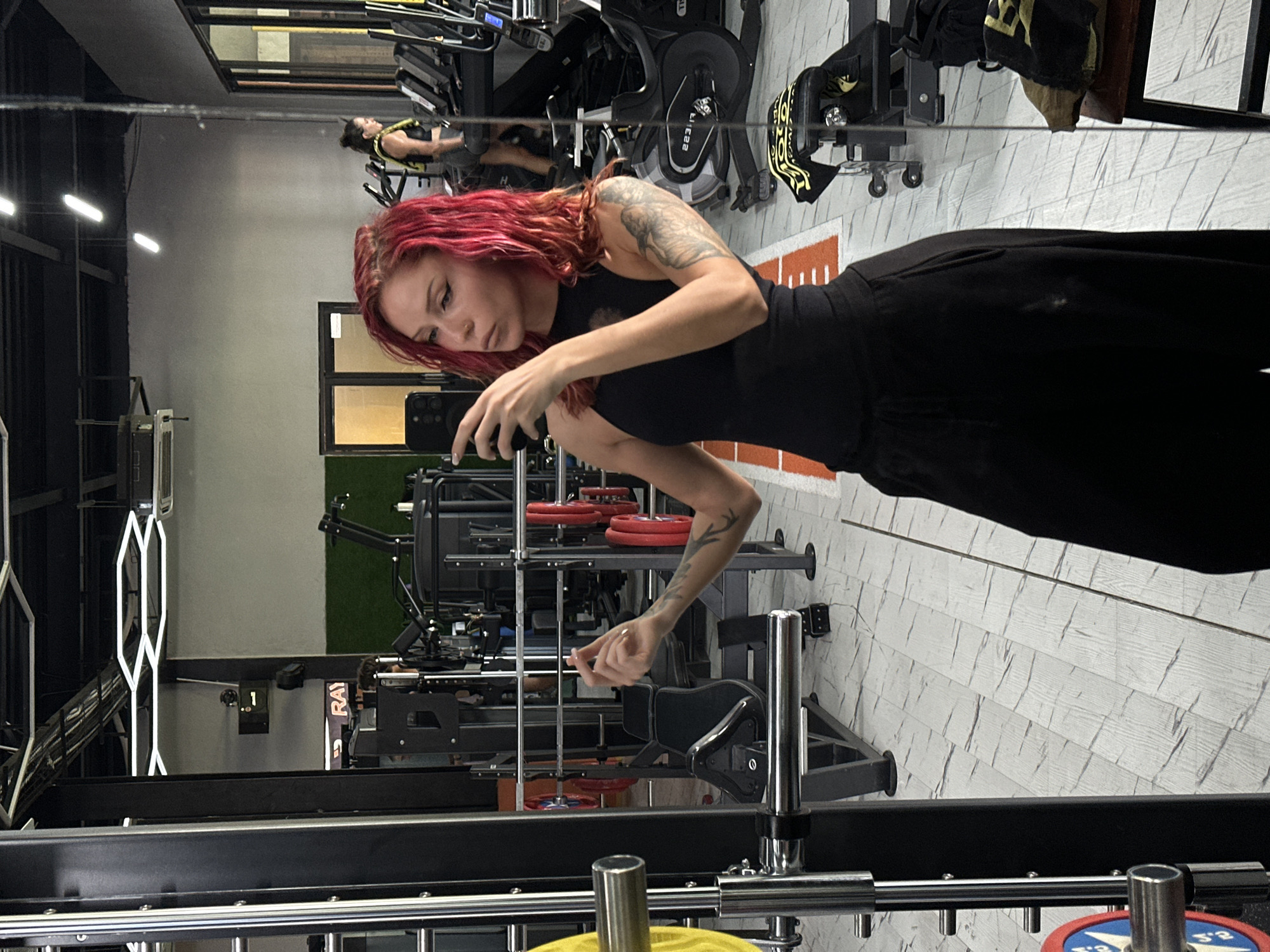A woman with red hair wearing a black tank top and black pants is taking a selfie in a gym. Gym equipment is visible in the background. The gym is located in North Kuta, Indonesia. Another person is exercising on a stationary bike in the background.