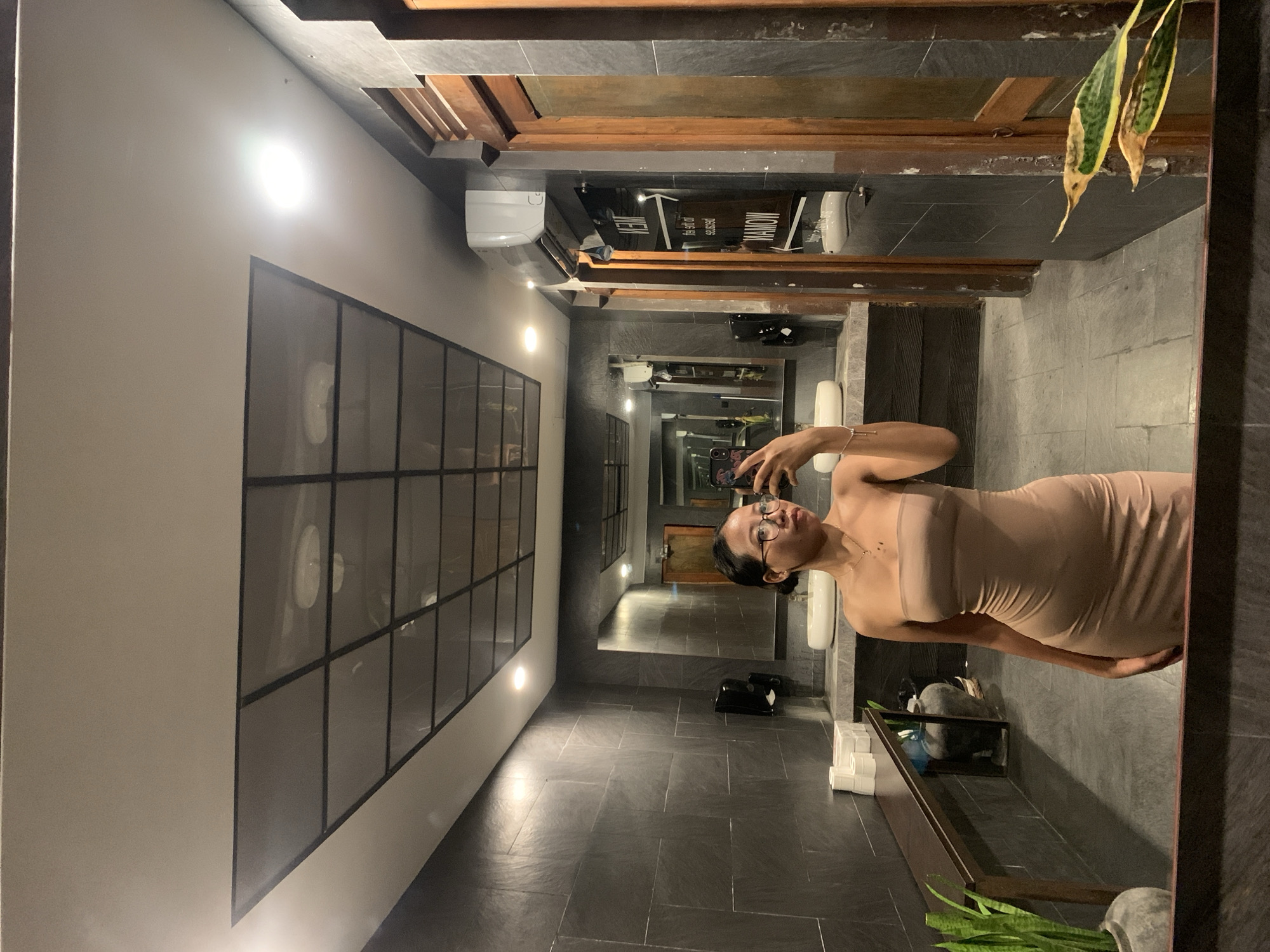 The image shows a woman taking a mirror selfie in a bathroom. The bathroom has gray stone floors and walls. A large window with black frames and white circular inserts is on the left side of the frame. The bathroom appears to be long and narrow, with a sink, toilet, and other bathroom fixtures visible in the mirror's reflection. The woman is wearing a nude-colored dress and glasses. The image was taken in North Kuta, Indonesia.