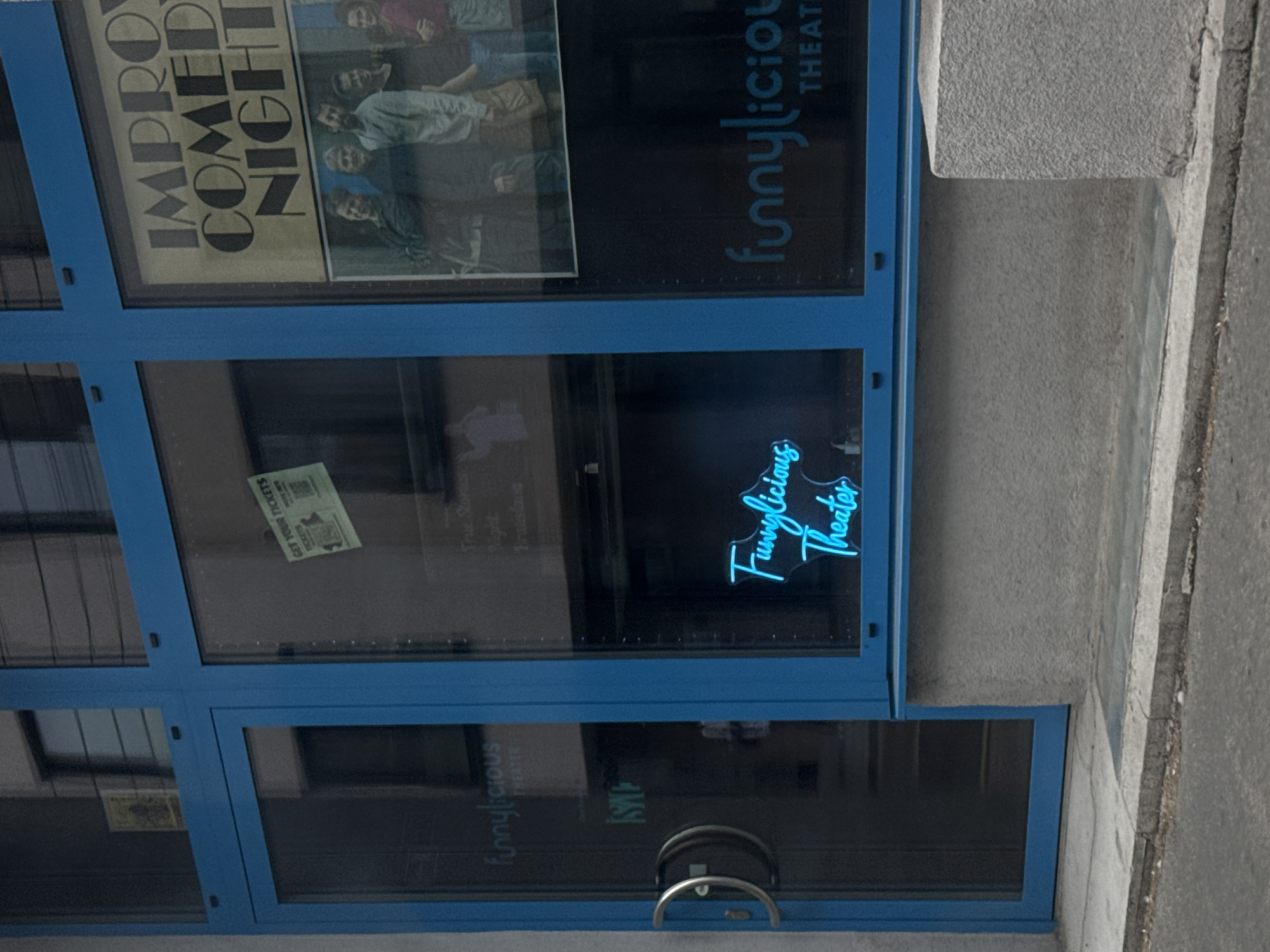 The image shows the exterior of a building with a blue-framed glass facade. On the left side, a glass door with two metallic handles is visible. To the right of the door, within the glass facade, is a neon sign in bright blue that reads "Funnylucious Theater". Further to the right, a poster is displayed, advertising "Improv Comedy Night". Below the poster, the text "funnylucious THEATER" is visible in white lettering. The building is situated in Bratislava, Slovakia, as indicated by the text on a green flyer with "GET YOUR TICKETS" written on it, placed on the glass facade. The facade appears to be at street level, with a concrete sidewalk and asphalt road in the foreground.