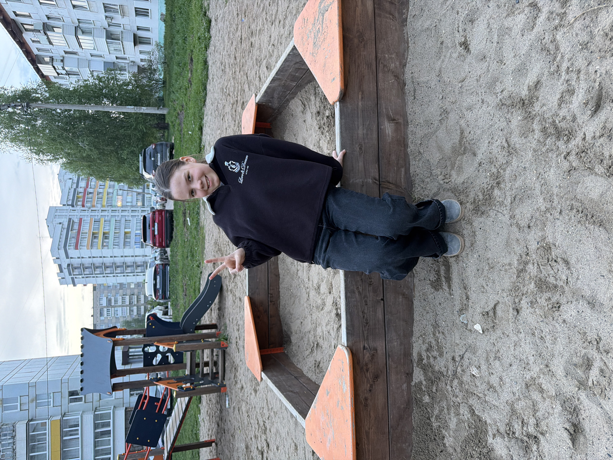 A young person is smiling and posing in a children’s playground in Chaikovsky, Russia. The individual is wearing a black sweatshirt and jeans and is standing beside a wooden sandbox structure, giving a “peace” sign with their left hand. The playground is situated within a courtyard, with residential buildings and parked cars visible in the background. Another playground structure with a slide can be seen in the lower left. The ground is covered with sand.