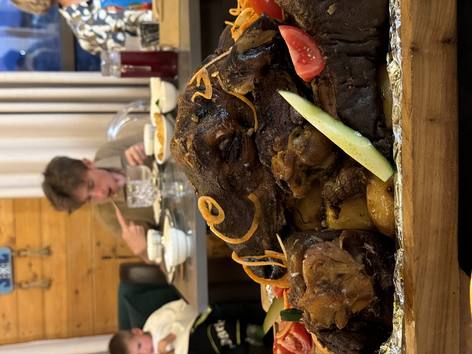 In this close-up, vertically oriented photograph, a large, dark, roasted meat dish, possibly a whole lamb or a large cut of beef, is the central focus. The meat is richly browned and appears to be seasoned with herbs and spices. It's garnished with a slice of tomato, a strip of cucumber, and thin, curly orange strands that might be fried onions or a type of vegetable crisp. The dish is presented on a wooden board lined with crinkled aluminum foil.
In the background, partially out of focus, a group of people are gathered around a table. One person, a young male with light brown hair, is seated and looking down at his hands, seemingly engaged in an activity at the table. Another individual, dressed in a patterned shirt, is visible further back. The setting appears to be an indoor dining area, with wooden paneled walls visible. The lighting suggests it could be evening or indoors with artificial lighting. The overall scene evokes a communal dining experience, likely a celebratory meal or a special occasion, given the prominent display of the roasted meat. There are no discernible weather cues or explicit location details beyond the general interior setting.