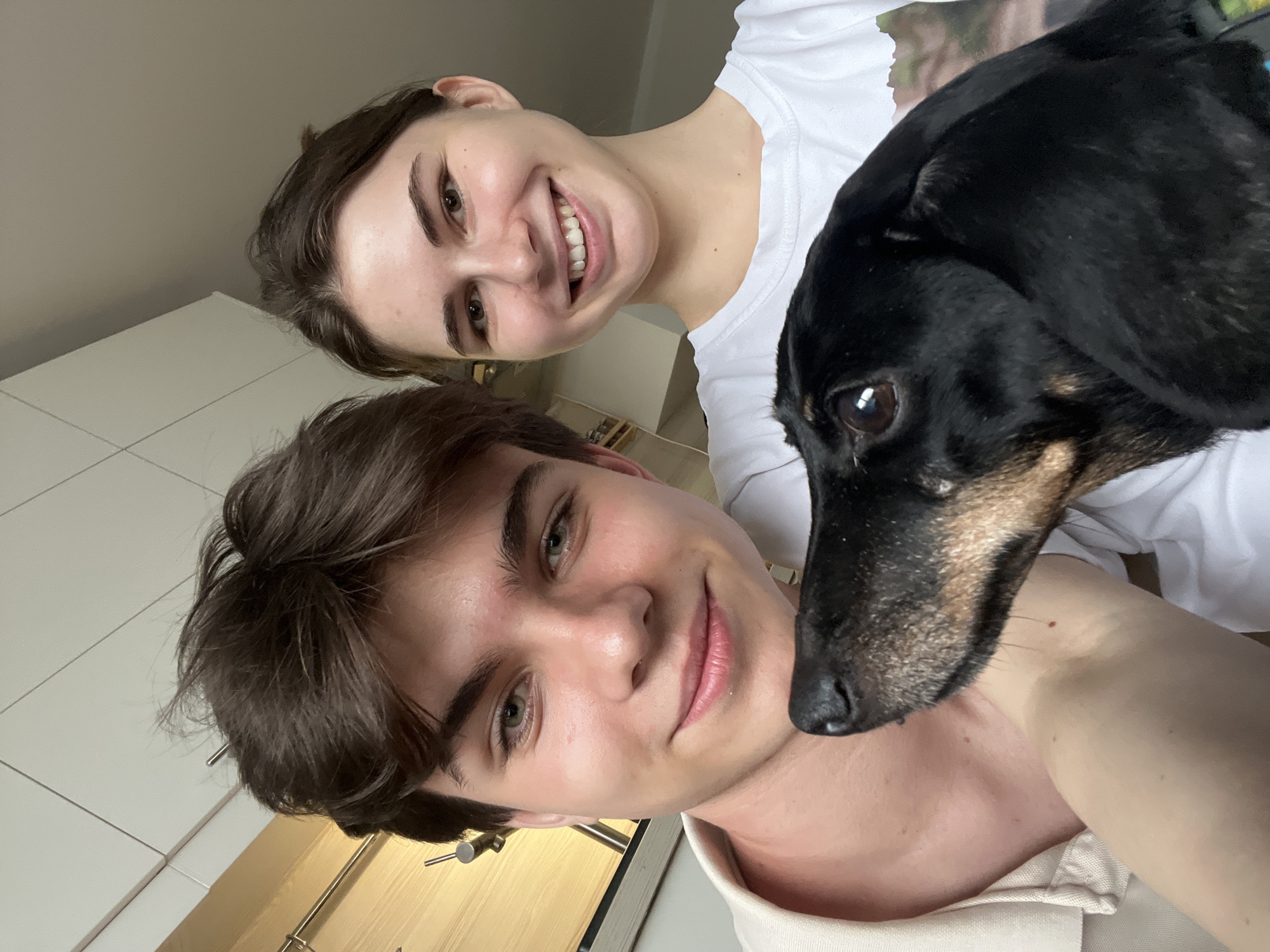 A selfie-style photograph captures two individuals, a male and a female, along with a dog. The location is Saint Petersburg, Russia. The male, positioned in the foreground, has brown hair, fair skin, and green eyes. He is wearing a light-colored shirt. The female, behind and to the left of the male, is smiling and wearing a white t-shirt. The dog, a black and tan dachshund, is positioned between the two people, its head near the male's neck. The background includes a white cabinet and a wooden countertop. The lighting appears to be natural, and the overall composition is casual.