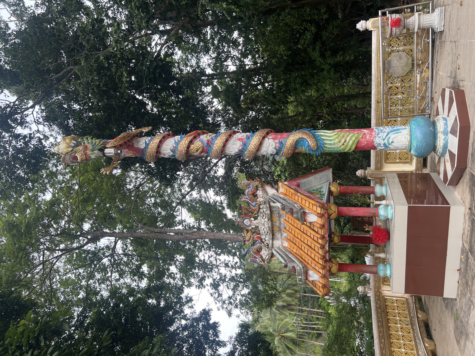 The image depicts a temple structure in Koh Phangan, Thailand. A tall, ornate pillar decorated with a coiled dragon sculpture stands prominently in the foreground. To the left, a small shrine with a multi-tiered, orange-tiled roof is visible, adorned with dragon figures along its ridge. The shrine contains an altar with offerings, including a red, floral-like decoration. The structures are situated outdoors, surrounded by lush green trees and foliage, suggesting a tropical environment. A decorative metal fence with gold accents runs along the perimeter of the temple grounds. The overall scene conveys a sense of cultural and religious significance within a natural setting.