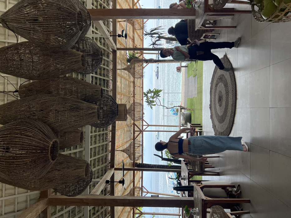 An outdoor restaurant with woven pendant lights above the seating area facing the ocean. A woman in a dark top and jeans stands near a table, and a man in a green shirt and dark pants poses on a round rug. Other patrons are seated at tables in the background. The restaurant has an open-air design with a view of the beach and ocean beyond.