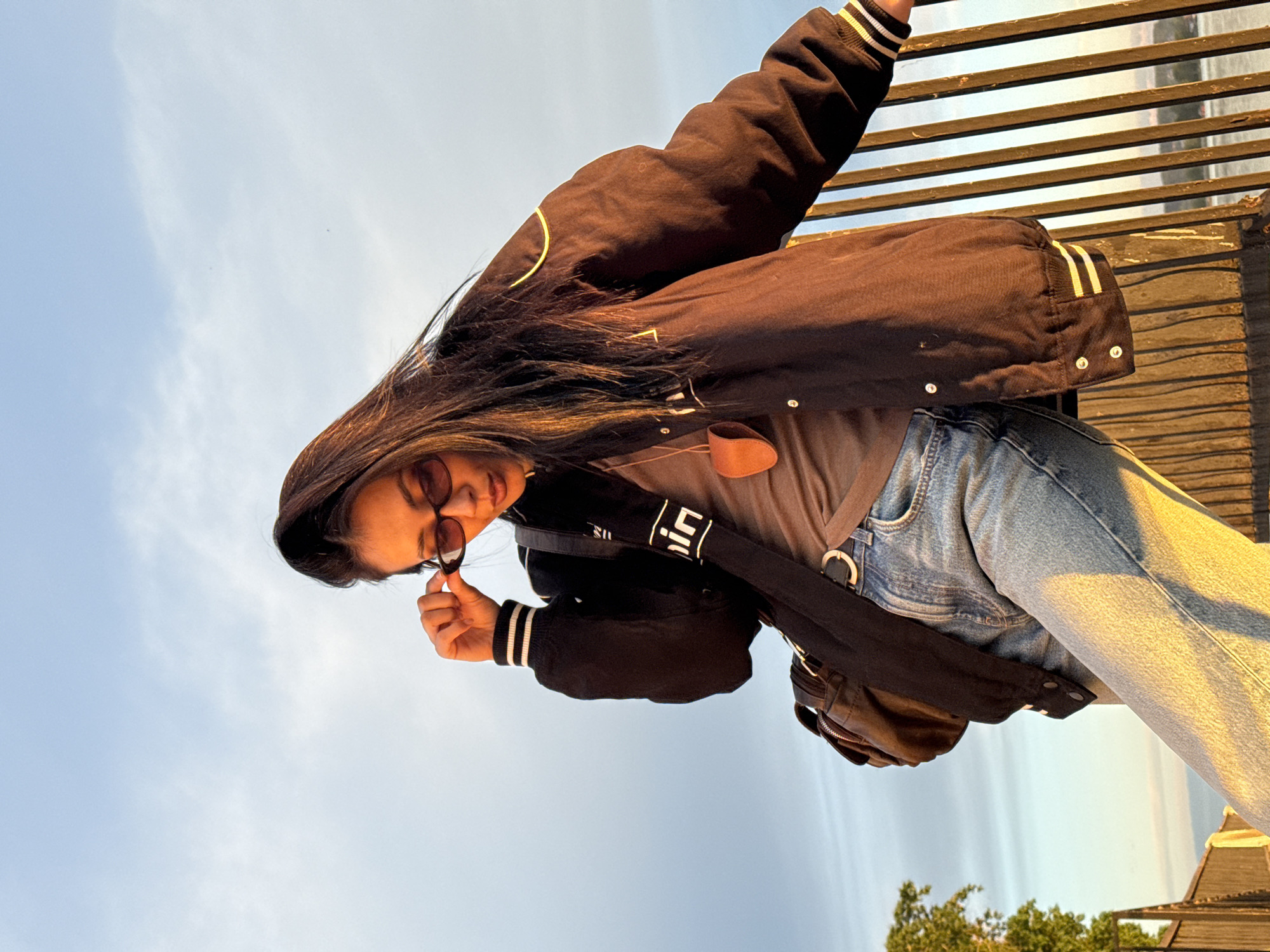 A young woman with long dark hair and sunglasses is positioned on an elevated wooden structure with horizontal slats, potentially a bridge or walkway. She is wearing a dark varsity jacket with white stripes on the cuffs, a grey t-shirt, light blue jeans, and a black bag slung across her body. She is holding the frame of her sunglasses with her left hand. The background consists of a light blue sky with some wispy clouds and a glimpse of greenery in the lower right corner. The image is taken from a low angle and the scene is illuminated by warm, possibly sunset lighting. There are no identifiable landmarks to confirm the exact location in Votkinsk, Russia.