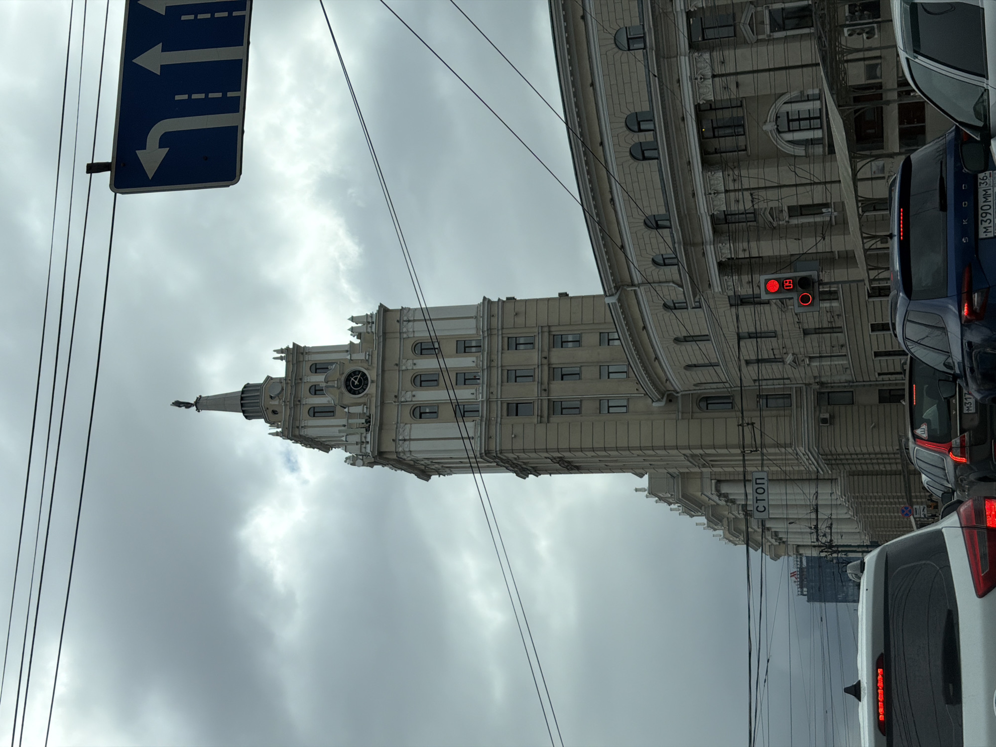 The image shows a street scene in an unknown city. A tall, light-colored building with a clock tower dominates the view. Power lines crisscross the sky in the foreground, and a blue street sign with directional arrows is visible on the left. A line of vehicles is stopped on the right side of the frame, with visible brake lights and license plates. A "STOP" sign is seen on a building facade in the background. The sky is overcast and cloudy.
