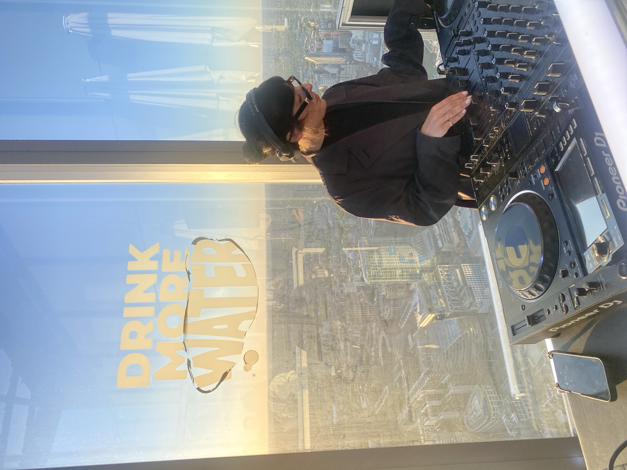 A rotated image captures a DJ performing high above a sprawling city, likely during the golden hour of sunrise or sunset. The primary subject is a person, seen from behind and slightly to the side, wearing headphones and glasses, with one hand on a professional DJ mixer. They are dressed in a dark jacket.
To the right, a set of professional DJ equipment is visible, prominently featuring a Pioneer DJ mixer and two Pioneer DJ media players. A smartphone rests on the surface next to the equipment.
The setting is a high-rise building, with a large window offering a panoramic view of a dense urban landscape, stretching out under a clear sky. The city below is illuminated by warm, soft light, indicative of either early morning or late afternoon.
Notably, the words "DRINK MORE WATER" are displayed in a light yellow-orange font on the window, styled with a water splash motif. Reflections of interior elements or distant structures are visible on the upper section of the window. The overall scene suggests a DJ set taking place in an elevated, urban environment, such as London.
