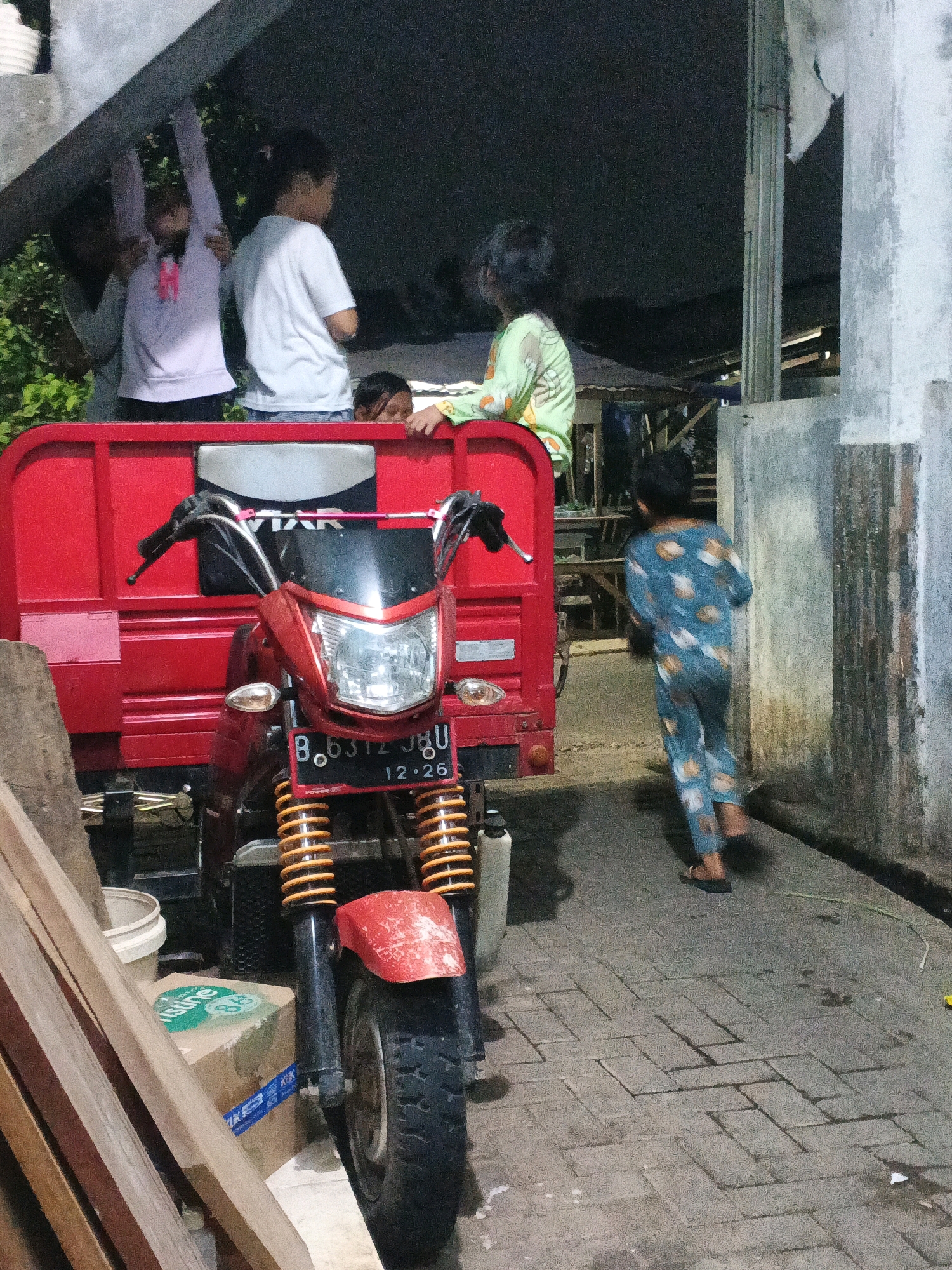 In Kadu, Indonesia, at night, a red tricycle-like vehicle is parked on a paved surface. A motorcycle is attached to the front of the tricycle. Several children are present. Three children are standing on top of the tricycle, one of whom is wearing a white t-shirt and blue shorts, another a light purple shirt with a pink ribbon, and the third a light green shirt with a pattern. Another child, wearing a blue pajama set with animal prints, is walking away from the tricycle towards a concrete wall. The environment appears to be an outdoor area with some debris and building structures visible.