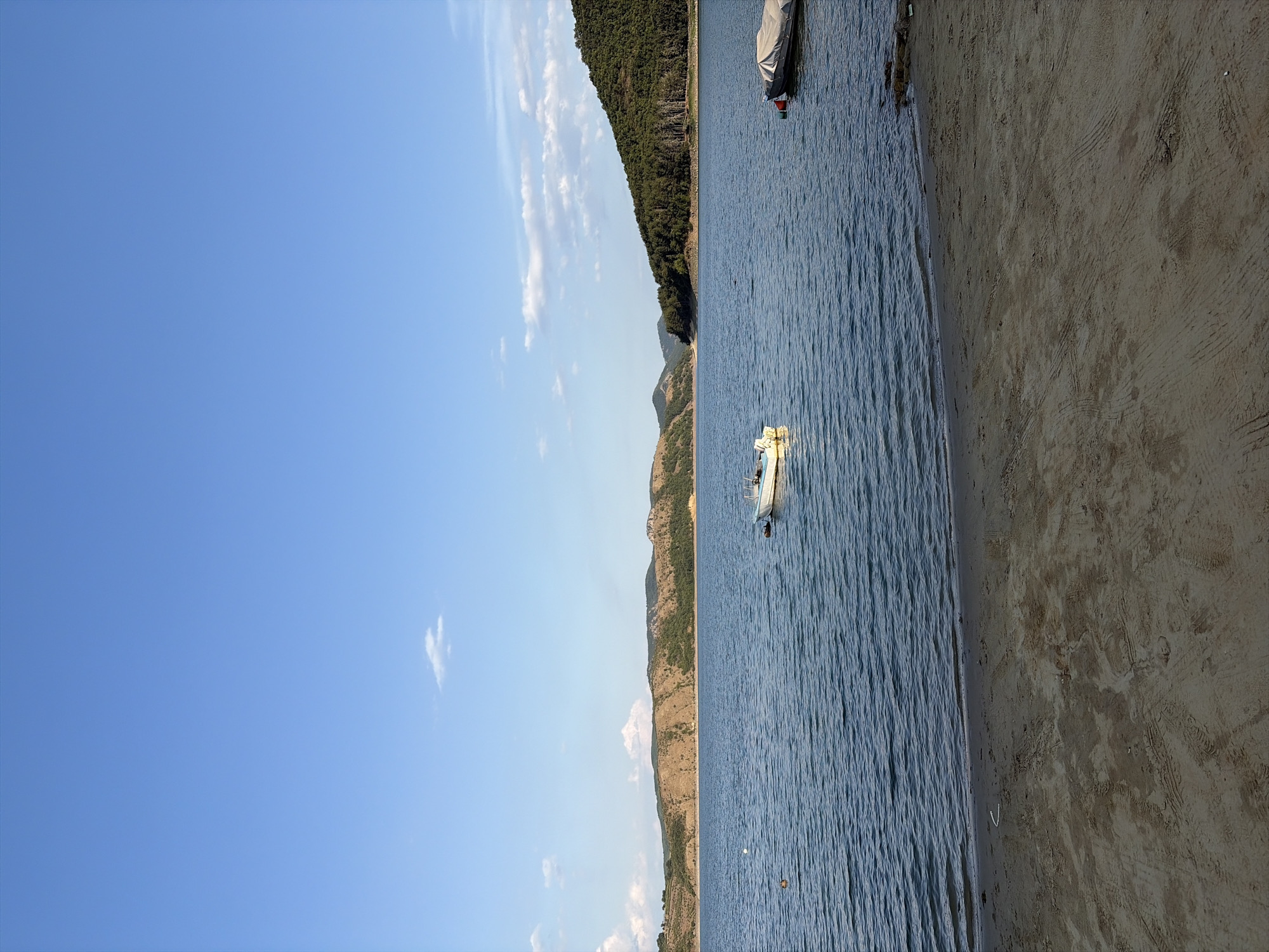 An eye-level, vertical, medium shot captures a serene lake scene under a bright blue sky dotted with wispy clouds. The foreground features a sandy beach with tire tracks. The middle ground is dominated by a wide expanse of calm blue water, rippled by a gentle breeze. Two small boats are visible; one is a white motorboat with a yellow trim, anchored in the center of the lake, and another is partially covered by a grey tarp, moored to the right. In the background, rolling hills covered with dry brush and scattered green trees rise from the water's edge, with more densely forested hills on the right. The overall atmosphere is peaceful and natural.