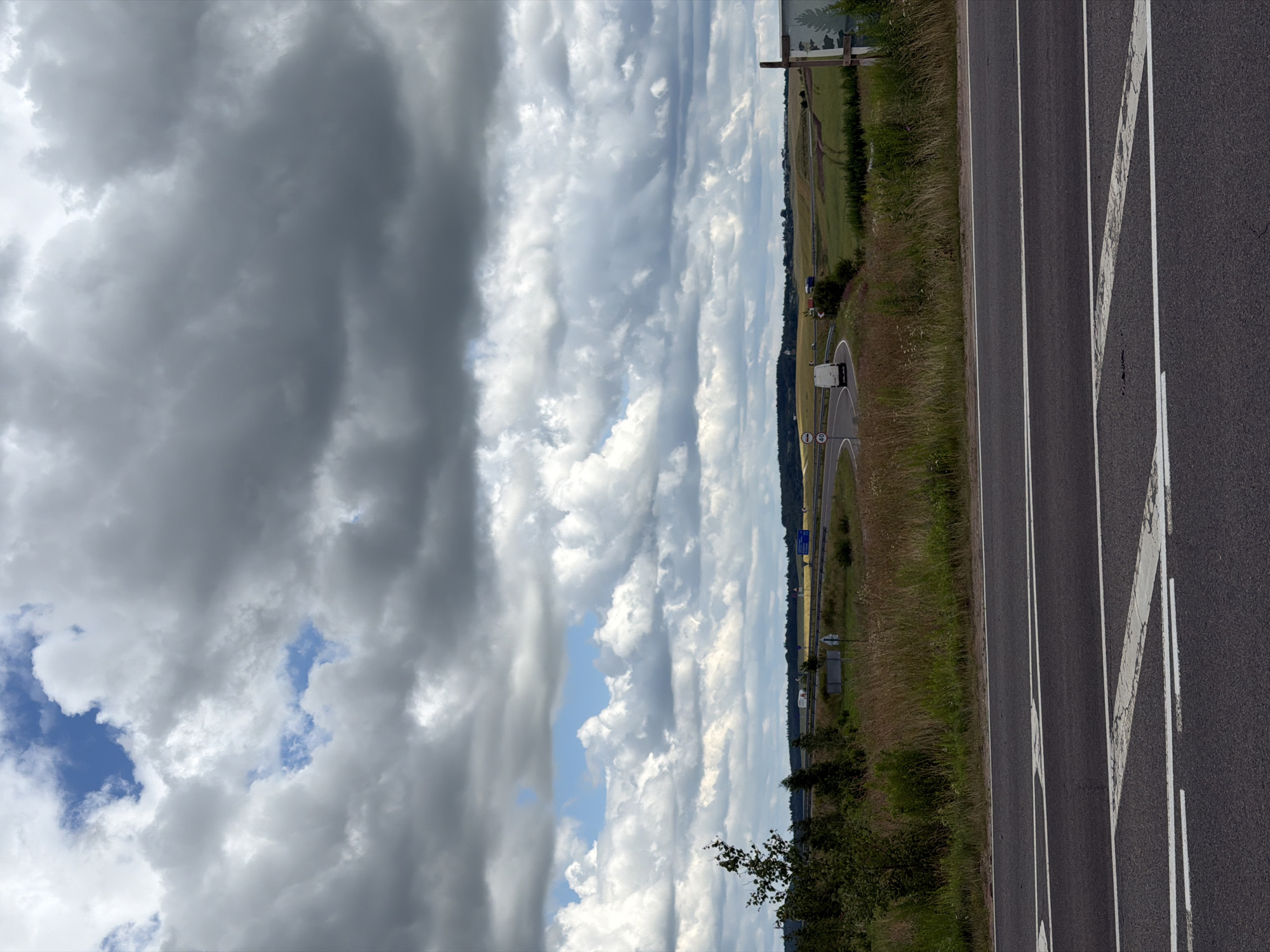 The image depicts a highway in the Папярнянскі сельскі Савет, Belarus, under a cloudy sky. The foreground features an asphalt road with white lane markings, including a chevron pattern. Beyond the road, a grassy embankment leads to a highway interchange. Several vehicles, including a white van and trucks, are visible on the highway. The surrounding landscape is characterized by fields, trees, and a distant treeline. The sky is filled with dramatic, dark clouds interspersed with patches of blue sky. A signpost with information is partially visible on the right side of the image.