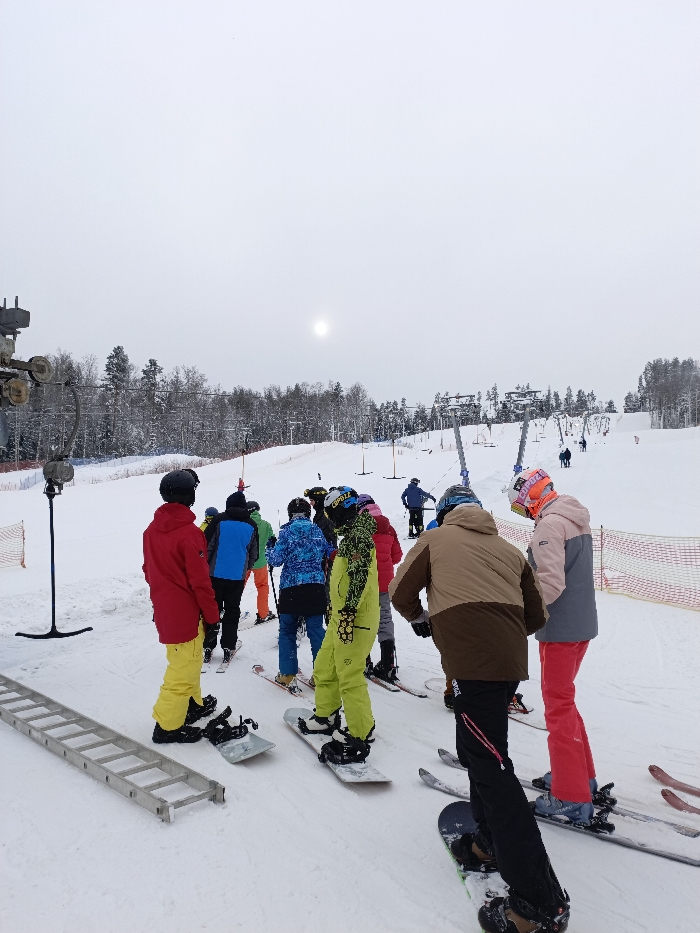 The image depicts a snowy ski slope at a resort in Svetloe, Russia, under an overcast sky with a visible hazy sun. In the foreground, a group of approximately ten individuals, dressed in various brightly colored ski and snowboard attire including helmets and goggles, are gathered on the snow. Most are facing uphill, appearing to be in a loose queue for a ski lift. A person in a red jacket and yellow pants stands on the left, near a snowboard lying flat. To their right, several individuals on skis are visible. In the center-right foreground, a person in lime green gear stands on a snowboard, while another in a brown and beige jacket is also on a snowboard. A person in a pink and grey jacket with red pants is on skis to the far right. A multi-section metal ladder lies flat on the snow in the lower-left. Partial ski lift infrastructure, including a support structure, is visible on the far left. In the midground, a wide snow-covered slope extends uphill, bordered by orange safety netting on the right. Multiple ski lift towers, consistent with T-bar or platter lifts, are seen further up the slope, with additional individuals visible using or approaching them. The background features a dense forest of snow-covered evergreen trees.