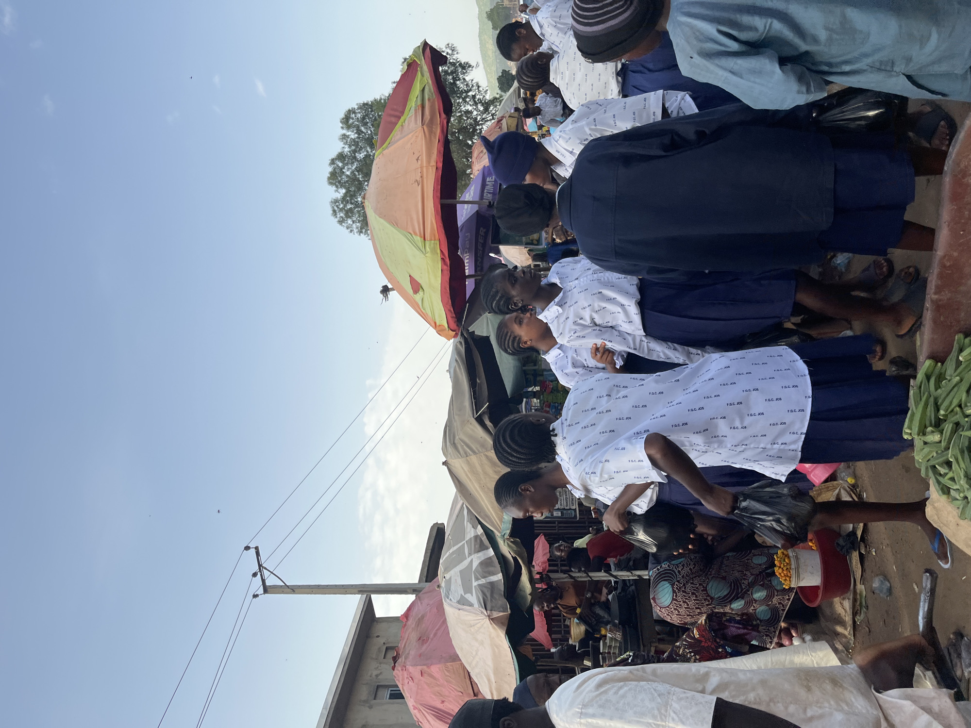 This media file captures a vibrant scene at a market in Rafin Par, Nigeria. The image is oriented vertically, giving a sense of depth and activity.
The primary subjects are a group of people, many of whom appear to be young women or girls, dressed in what seem to be school uniforms – white shirts with repeating text and dark skirts or trousers. They are clustered around market stalls, some looking at goods, others engaged in conversation. One woman in the foreground is wearing a colorful patterned dress. Men are also present, some in traditional attire.
Objects within the scene include various market stalls covered with umbrellas and tarps in muted colors like red, yellow, and green. Bags, some black and some red, are visible, along with what appears to be produce such as okra and small yellow fruits in a red container. A utility pole with wires is present in the upper portion of the image, indicating an urban or semi-urban setting.
The activity appears to be everyday commerce at a local market. The time of day is likely daytime, given the bright sky and shadows. The weather seems clear and sunny. The expressions on the faces of the people are mostly neutral or engaged in their surroundings, suggesting a typical market day.
Visible text includes repeated instances of "F.G.C. JOS" on the white shirts, likely an acronym for a school or organization. Other text, such as "ANSFER" and "BIRTIME," are also present on banners or signs within the market, though their context is unclear.